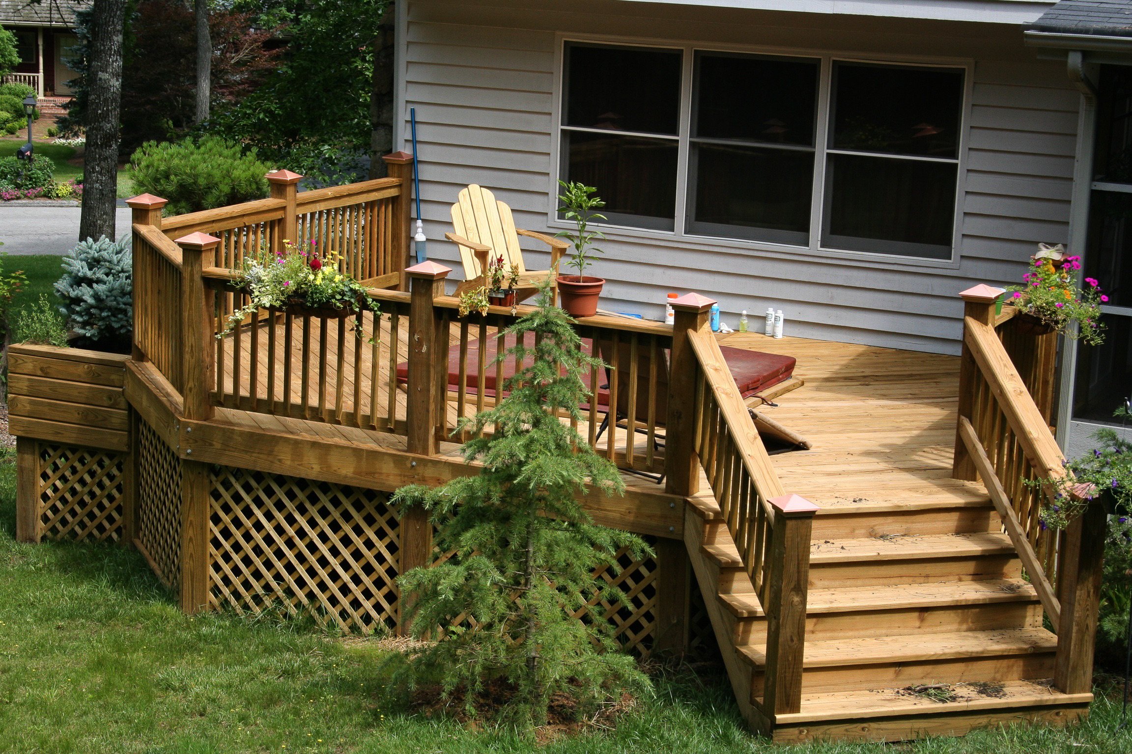 A wooden backyard deck with stairs leading down to a grassy yard, decorated with potted plants and outdoor furniture, attached to a house with gray siding and large windows.