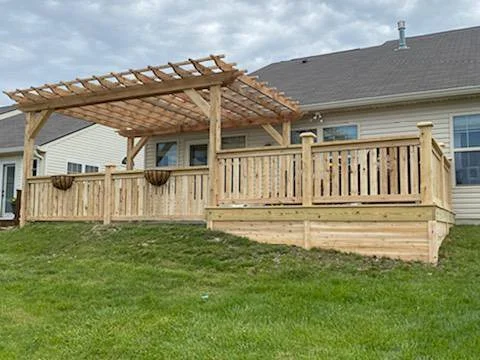 Wooden deck with a pergola, attached to a house, on a grassy lawn.