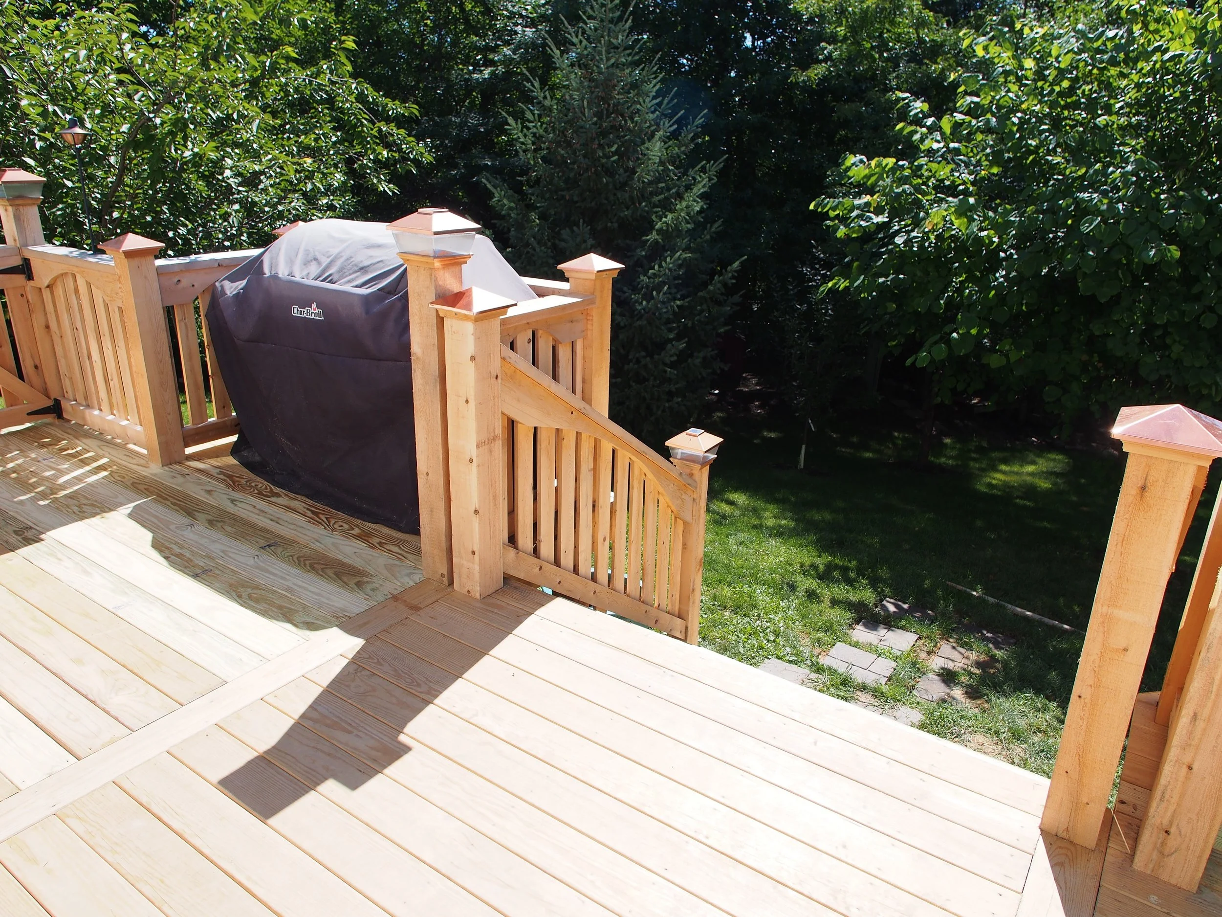 Wooden deck with a black covered grill on the left, surrounded by a wooden railing. In the background are lush green trees and grass.