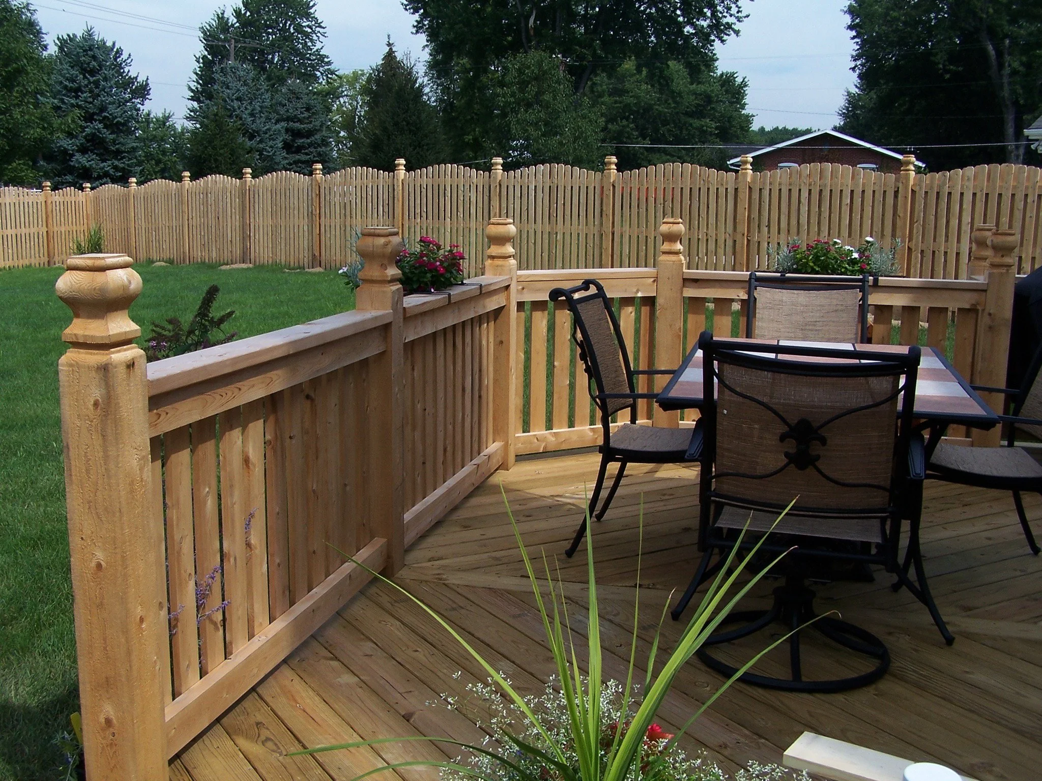 A wooden backyard deck with outdoor dining furniture and potted flowers, enclosed by a wooden fence, overlooking a green lawn and trees.