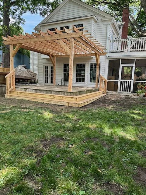 A house with a newly built wooden deck and pergola in the backyard, with grass, trees, and a grill covered with a tarp.
