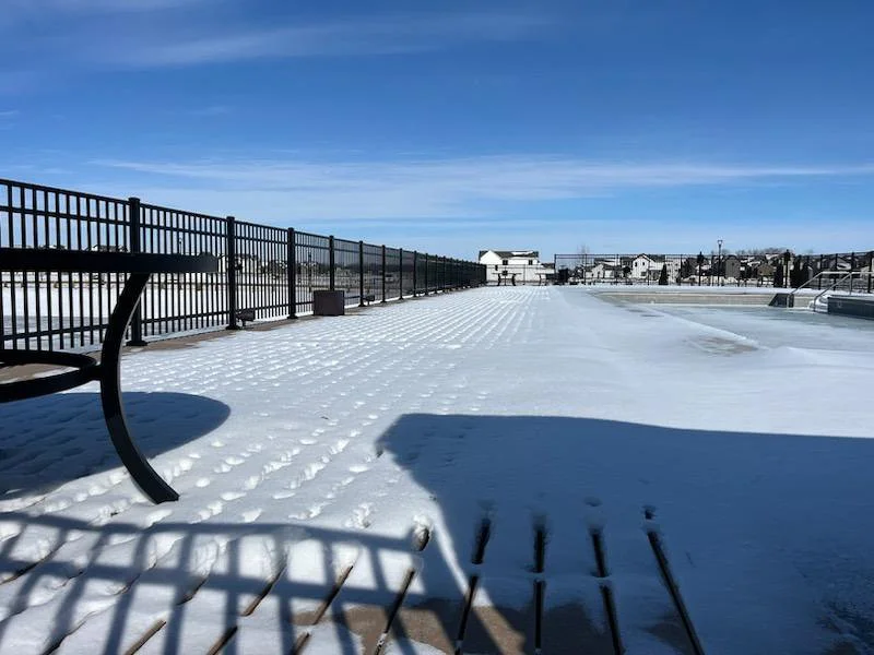 Snow-covered outdoor patio with a black metal fence, a bench, and distant buildings under a clear blue sky.