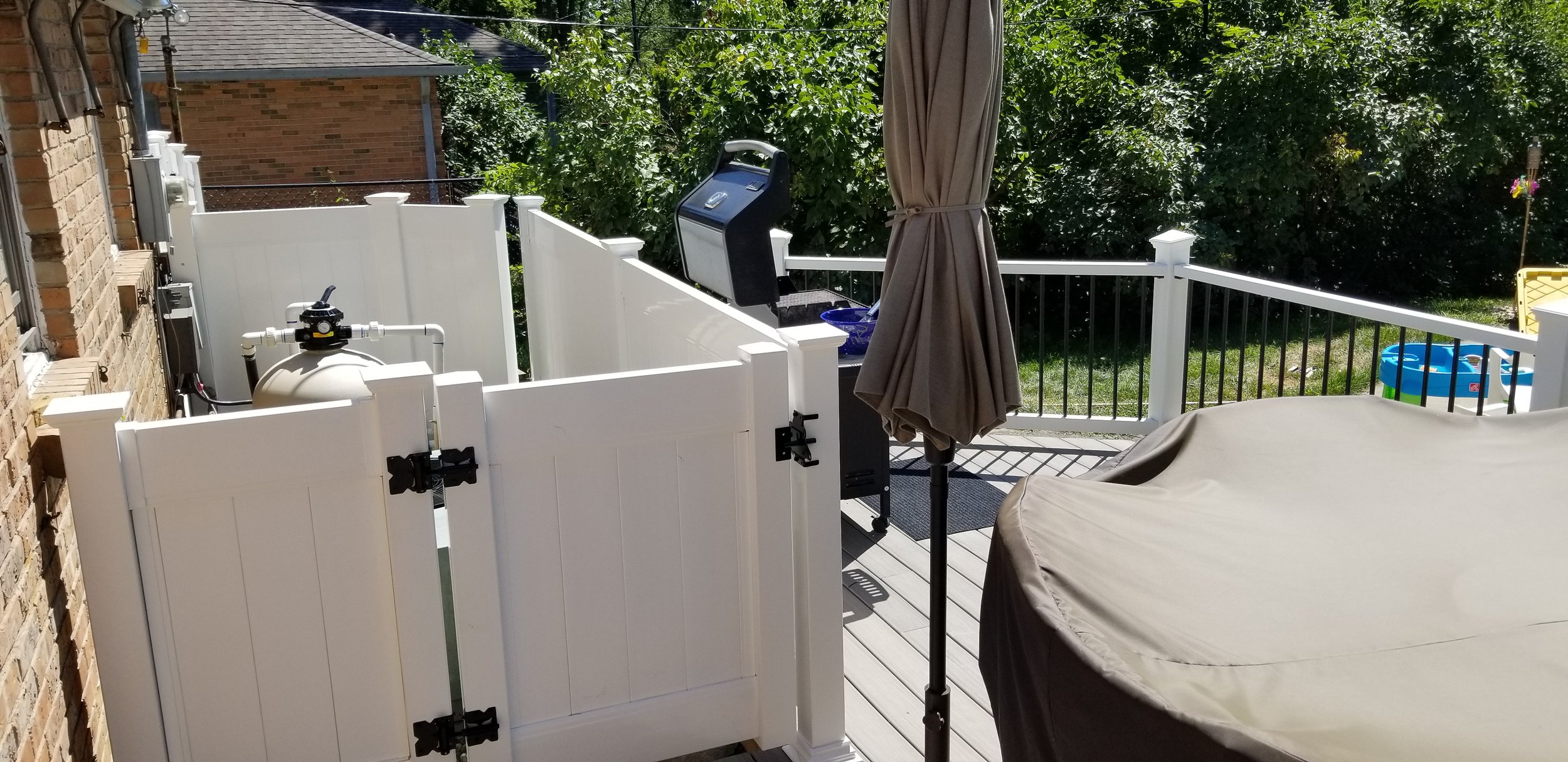 A backyard patio with a white fence, an outdoor grill, a closed patio umbrella, a covered piece of furniture, a small plastic pool, and lush green trees in the background.