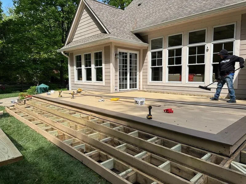 Construction workers installing a new wooden deck outside a house with beige siding and large windows, surrounded by green trees.