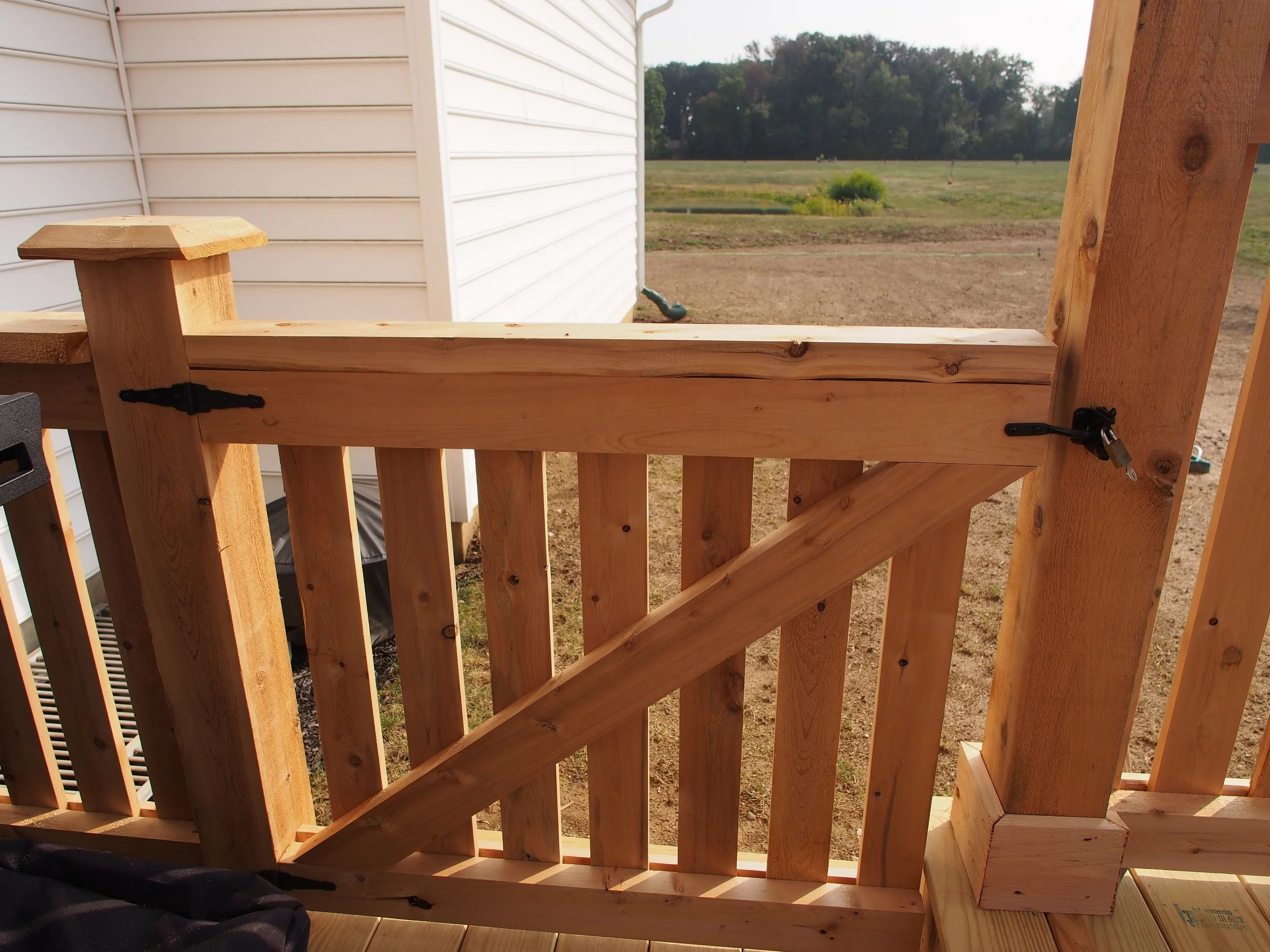 Close-up of a wooden railing with painted black hinges, attached to a partially built outdoor deck, overlooking a field and trees in the background.