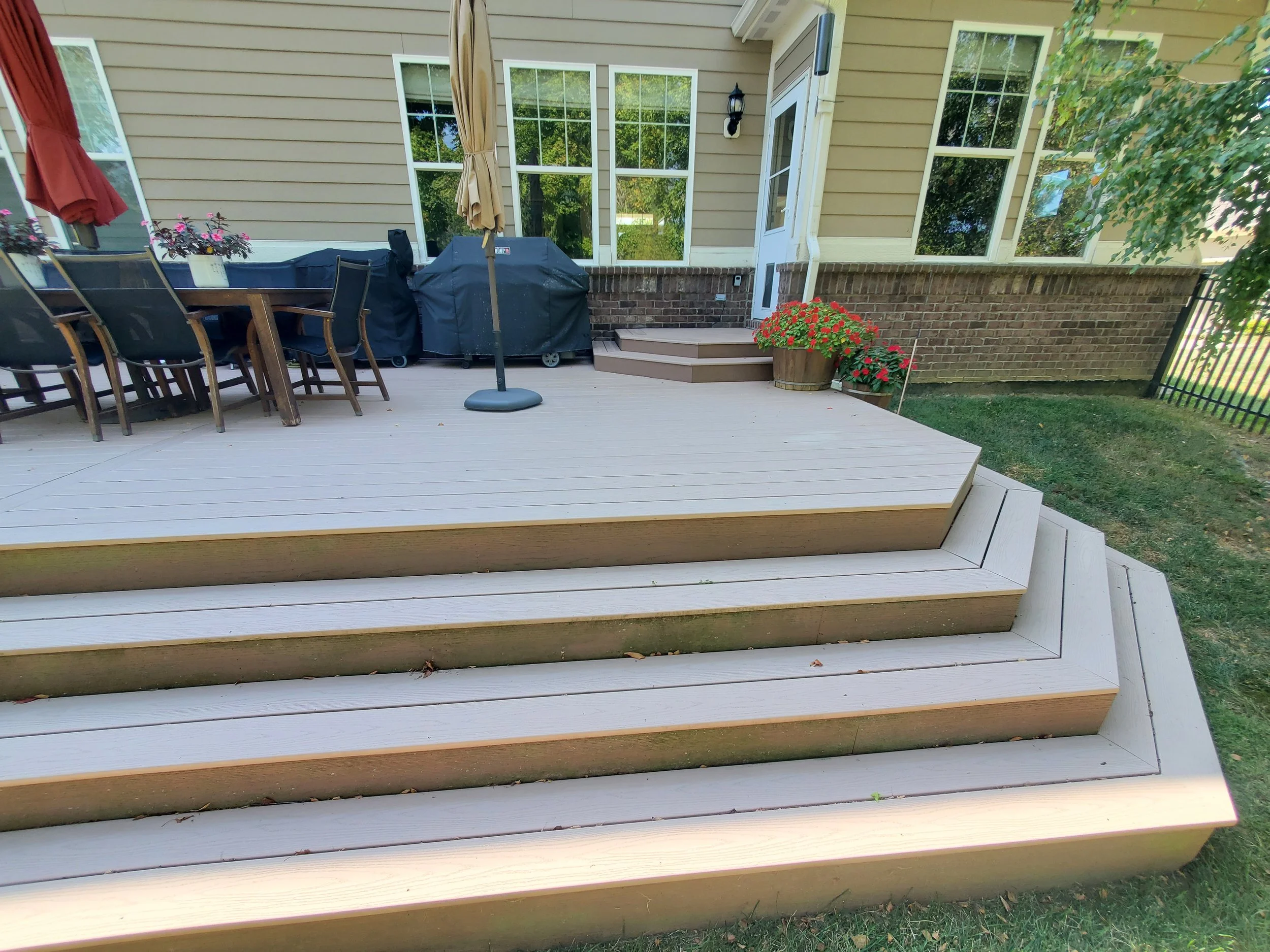 A backyard deck made of light-colored wood with stairs leading up to it, next to a grassy lawn. There is a patio table with chairs, a covered grill, potted flowers, and a house with yellow siding and multiple windows.
