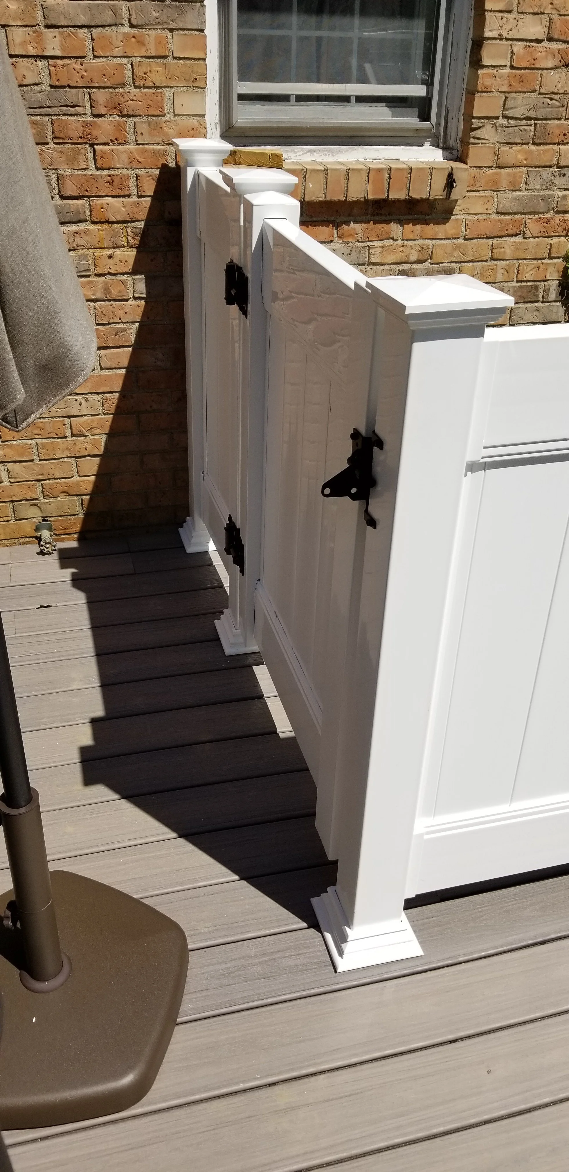 White wooden fence panel with black hinges on a wooden deck outside a brick house with a window.