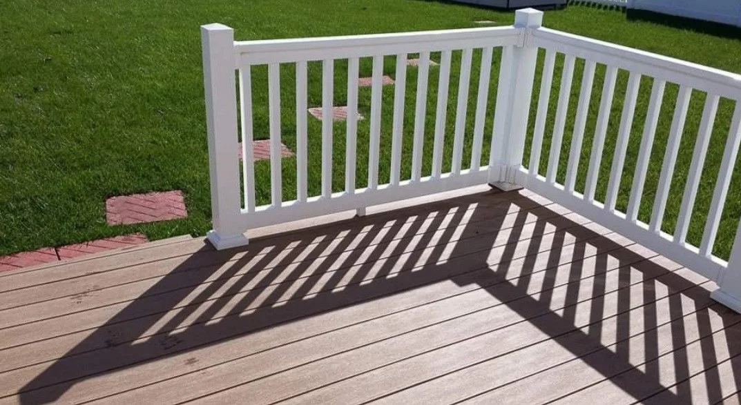 View of a white wooden deck railing casting shadows on the wooden deck, with a lawn and brick pathway in the background.