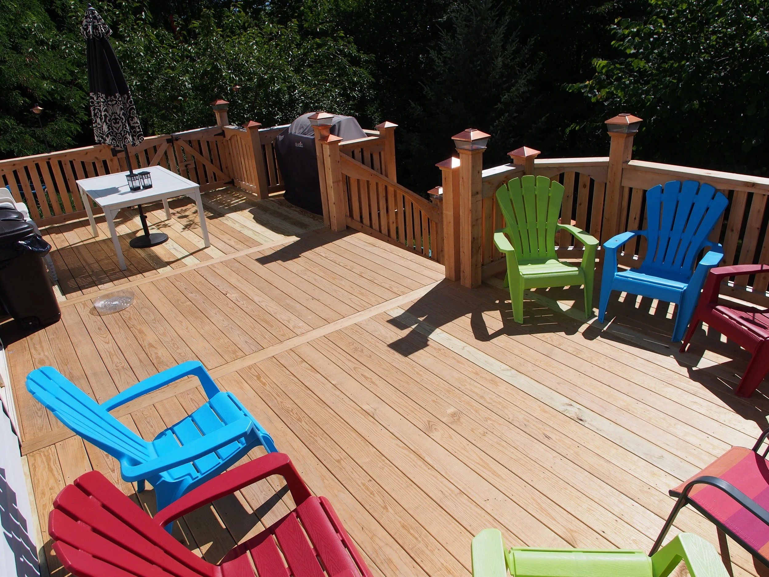 Colorful outdoor deck with several Adirondack chairs in red, blue, green, and pink, a white table with an umbrella, a black grill, and surrounded by trees.