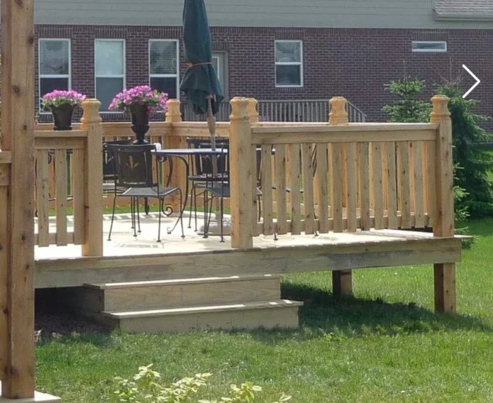 Newly built wooden deck with outdoor furniture and potted flowers, attached to a residential house, with steps leading down to a grass yard.