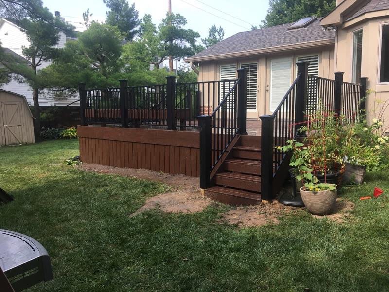 Wooden backyard deck with black railing and stairs, surrounded by green lawn, potted plants, and a house in the background.