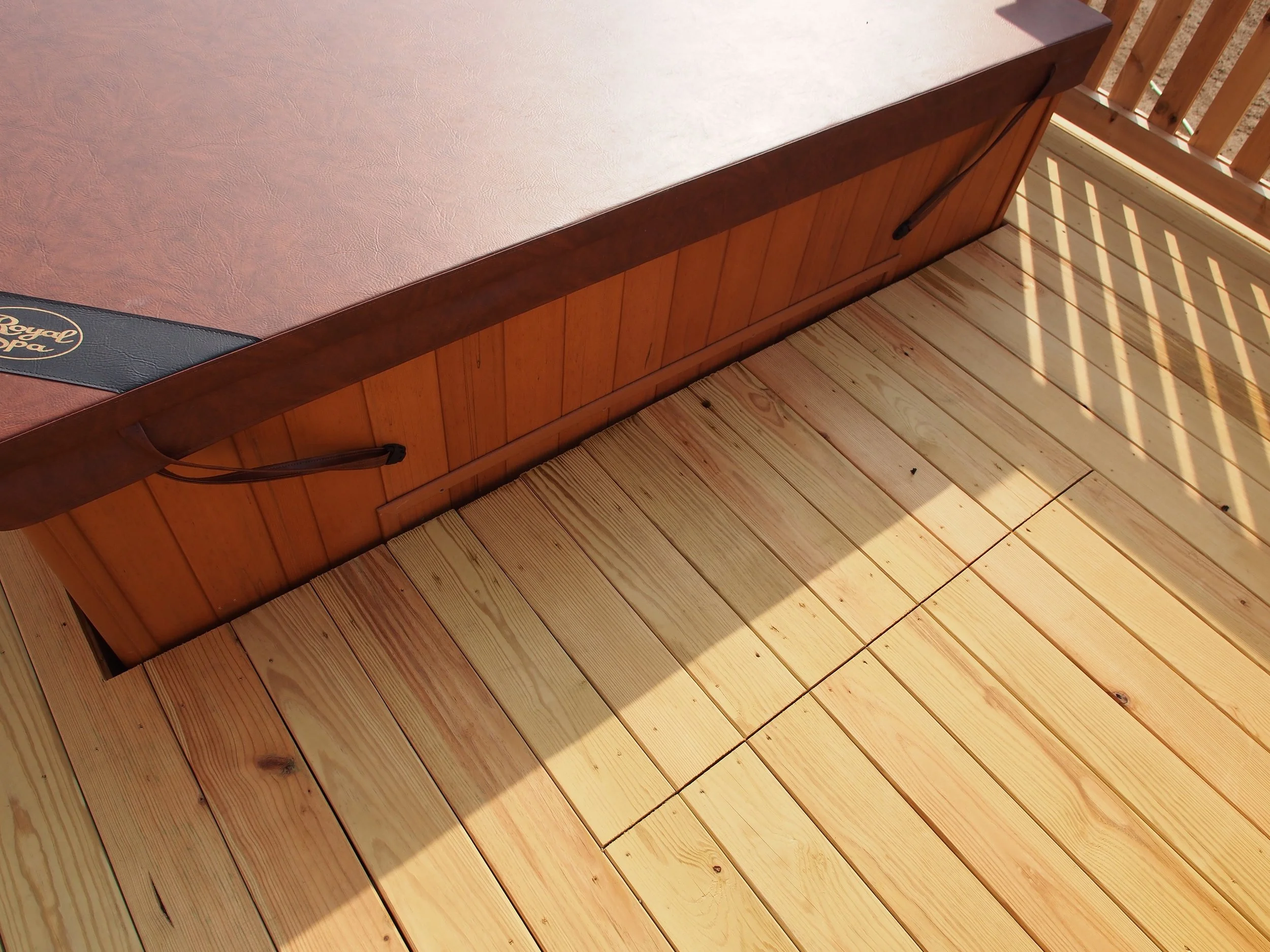 Close-up view of a wooden porch with a partially opened wooden hatch or trap door, revealing a well-lit interior space. The porch includes light-colored wooden planks, and a table with a brown surface and a black leather edge with a logo.