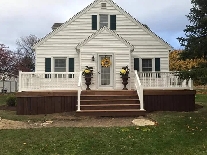 Front view of a white house with a large wooden porch decorated with yellow flowers and a wreath, set on a lawn with trees in the background.