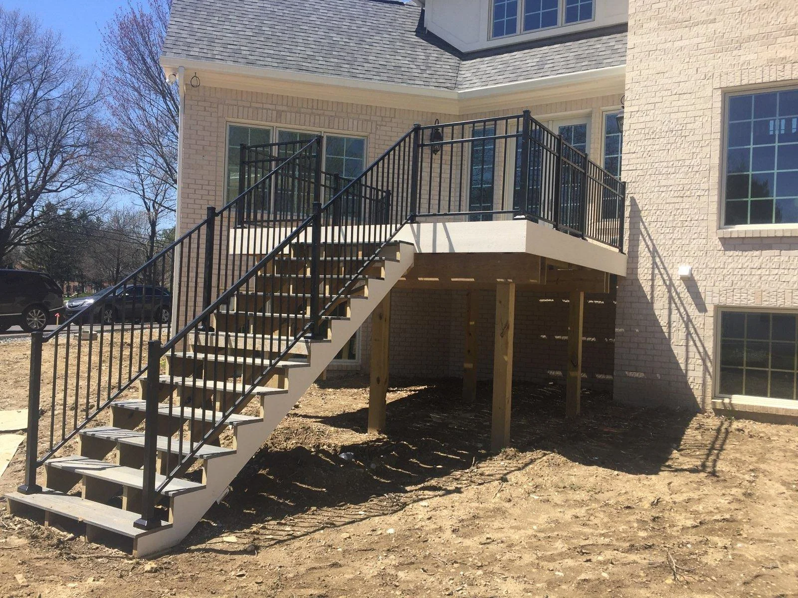 Newly built outdoor deck with stairs and black metal railing attached to a brick house, with a dirt ground underneath.