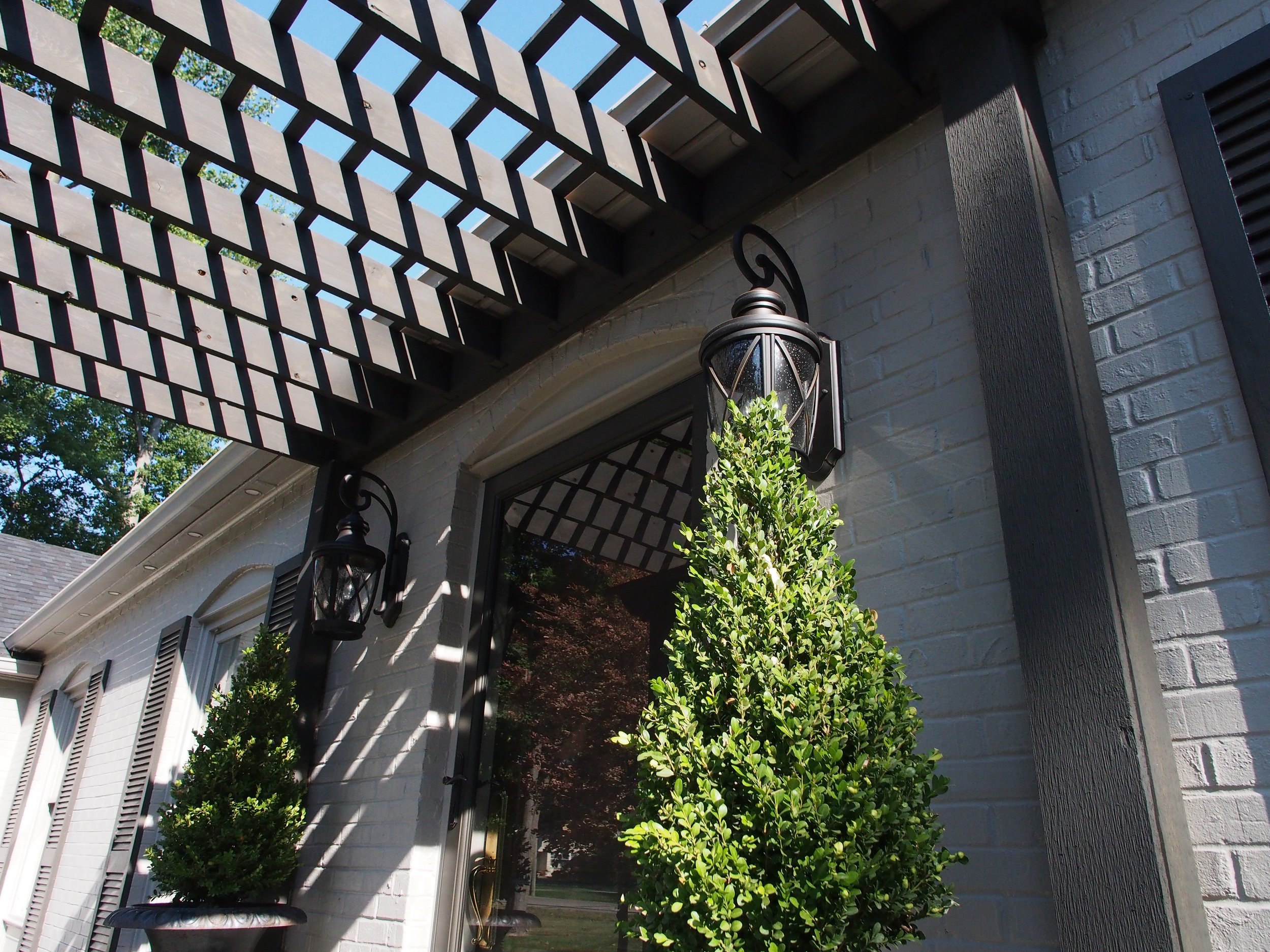 Outside view of a house showing a white brick wall, black shutters, hanging lantern lights, and two potted topiary plants on either side of the door, with a black pergola overhead casting shadows.
