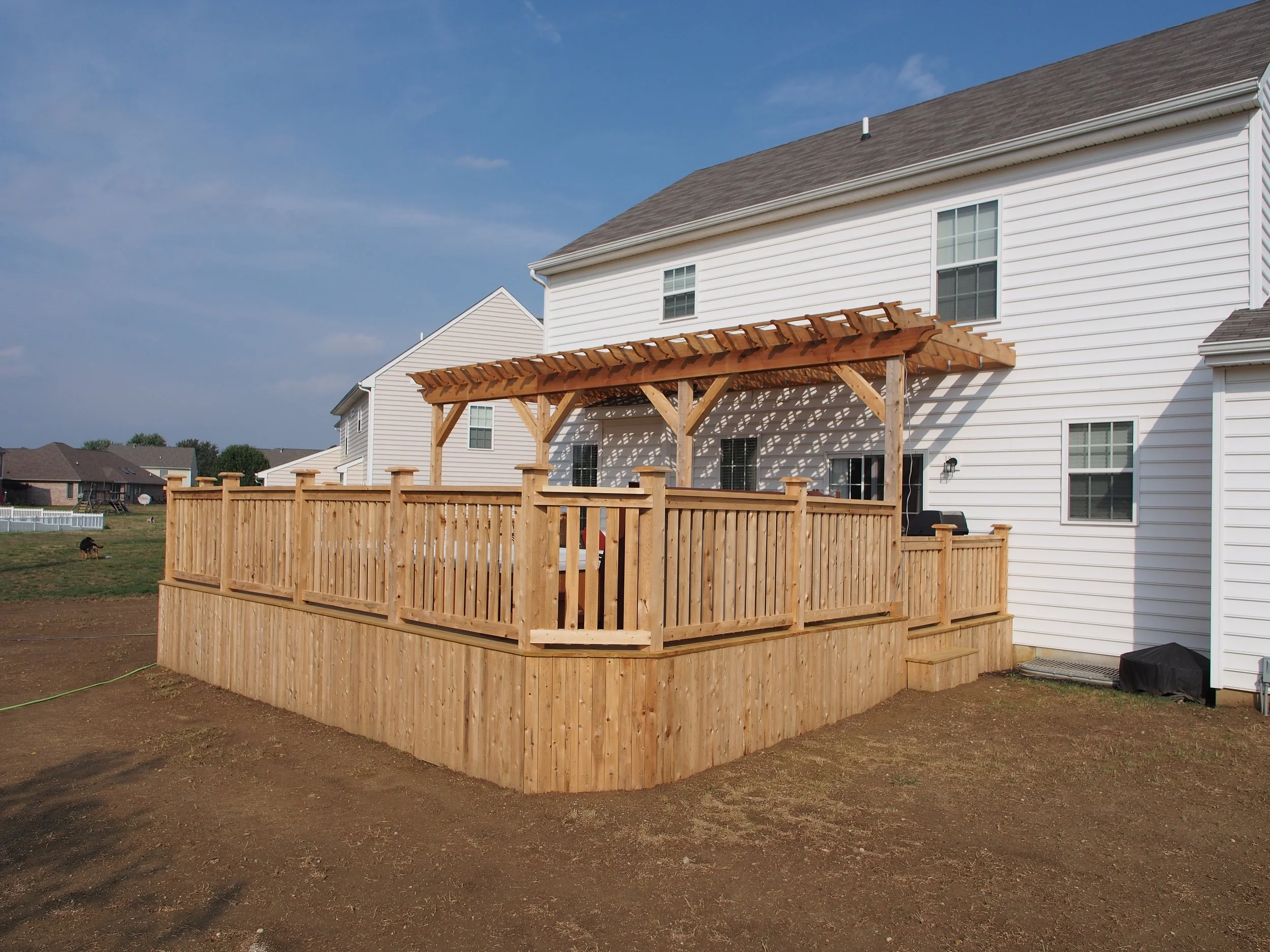 Newly built wooden deck with railing and pergola attached to the back of a white house.