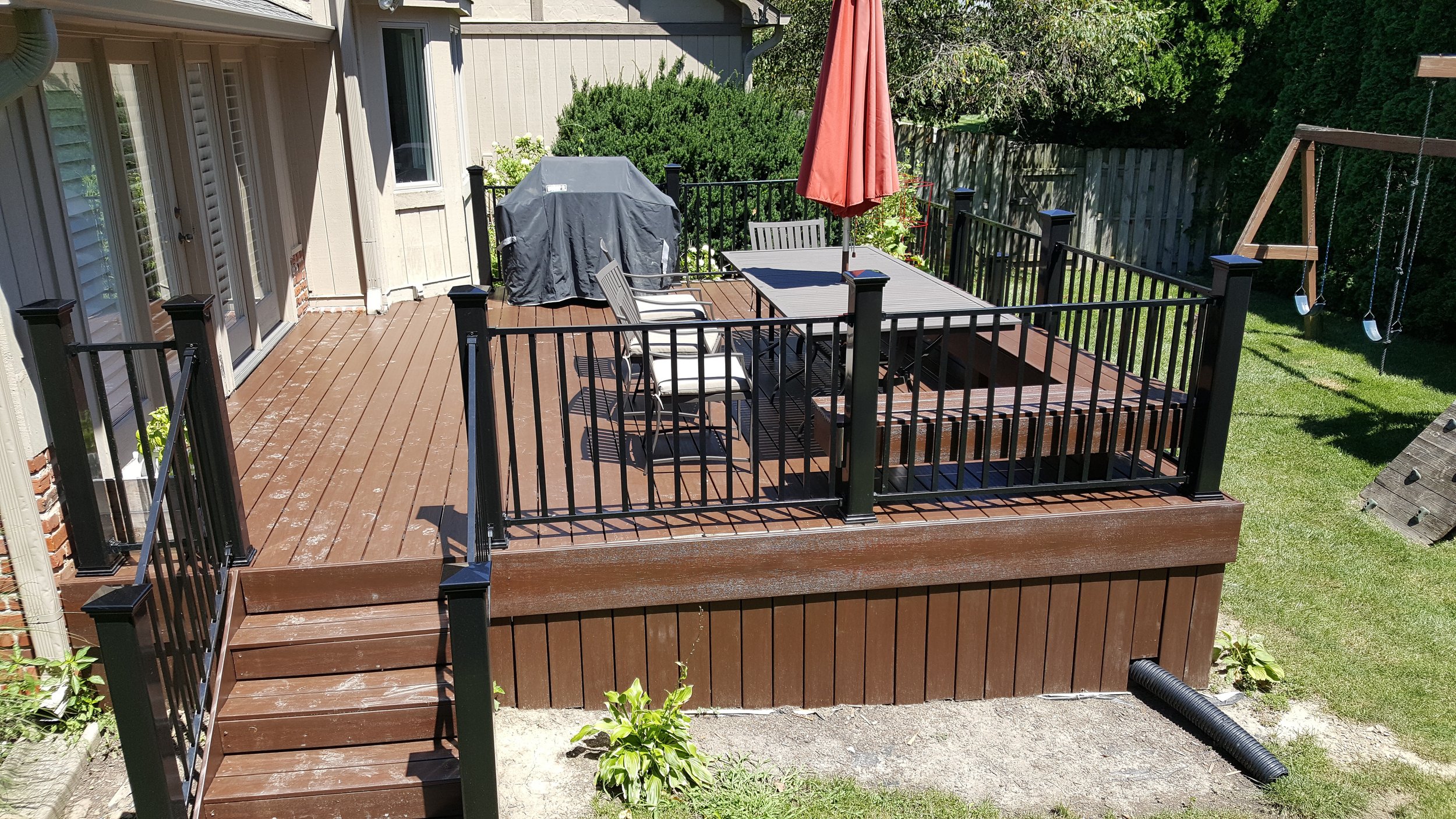 A wooden backyard deck with black metal railing, chairs, a table with an umbrella, a covered grill, a swing set with two swings, and various plants and greenery around.