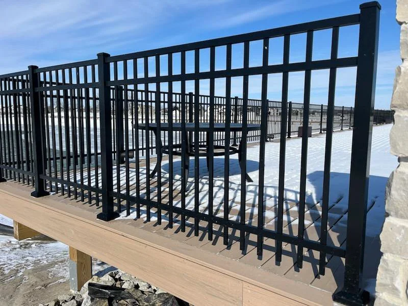 Black metal railing enclosing a wooden deck with a round outdoor table and chairs, snow on the deck, and a clear blue sky in the background.