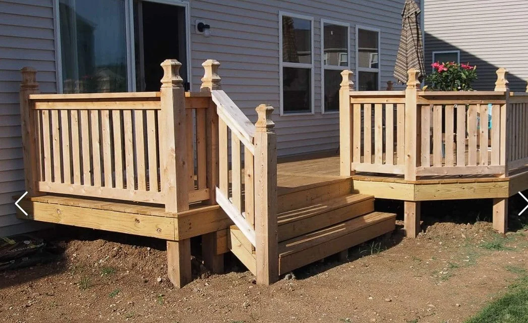 Newly built wooden deck attached to a house with stairs and a small railing, decorated with potted flowers and an umbrella on a sunny day.