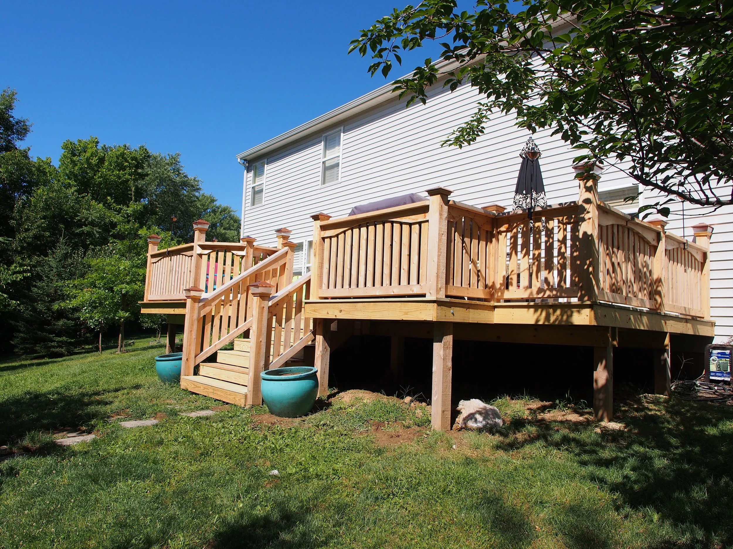 Backyard deck under construction with new wooden railings and stairs, surrounded by grass and trees, with a dog lying underneath, a white house with siding in the background, and a blue sky.