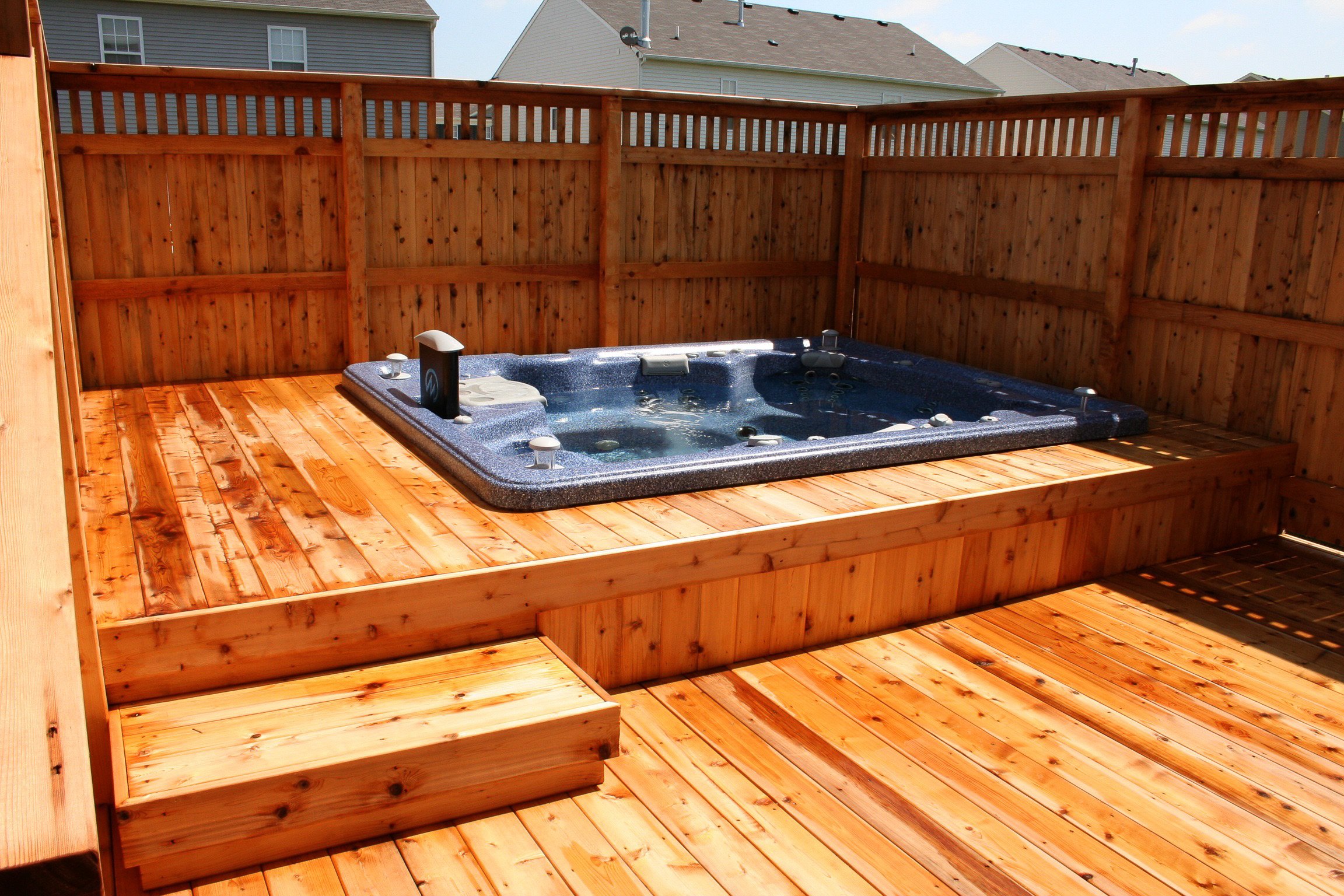A wooden deck with a hot tub surrounded by a wooden privacy fence, in a suburban backyard with neighboring houses in the background.