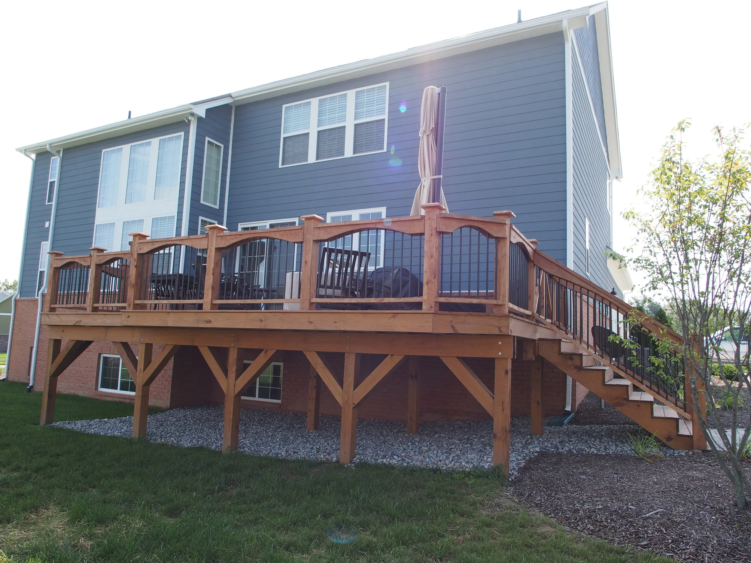A blue multi-story house with a large wooden deck and black railings. The deck has outdoor furniture and an umbrella, with stairs leading down to a grassy yard.