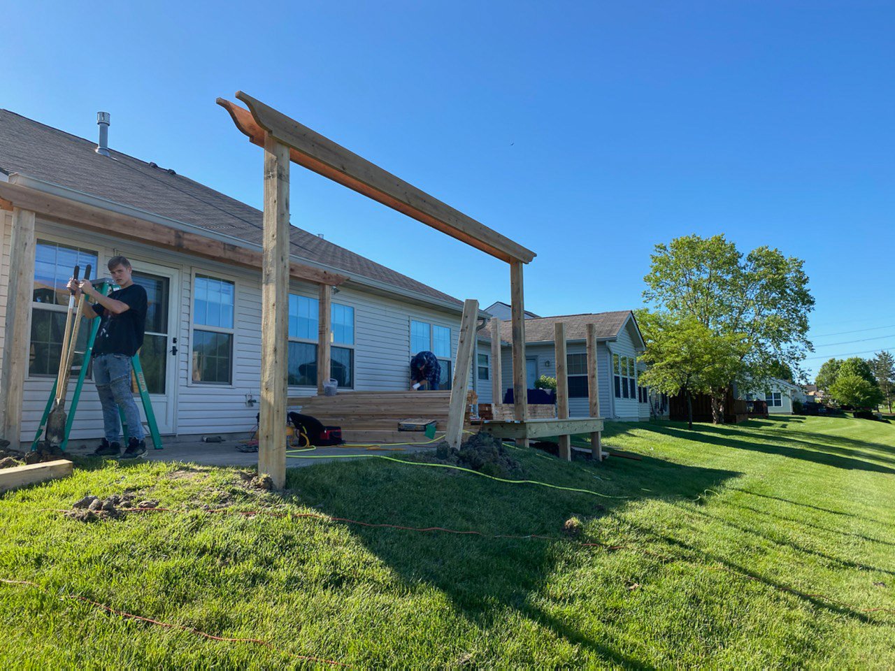 Two people are building a wooden deck outside a house. One person is on a ladder, holding a piece of wood, while the other is kneeling on the deck. The house has beige siding and large windows, and the area is surrounded by green lawn and trees.