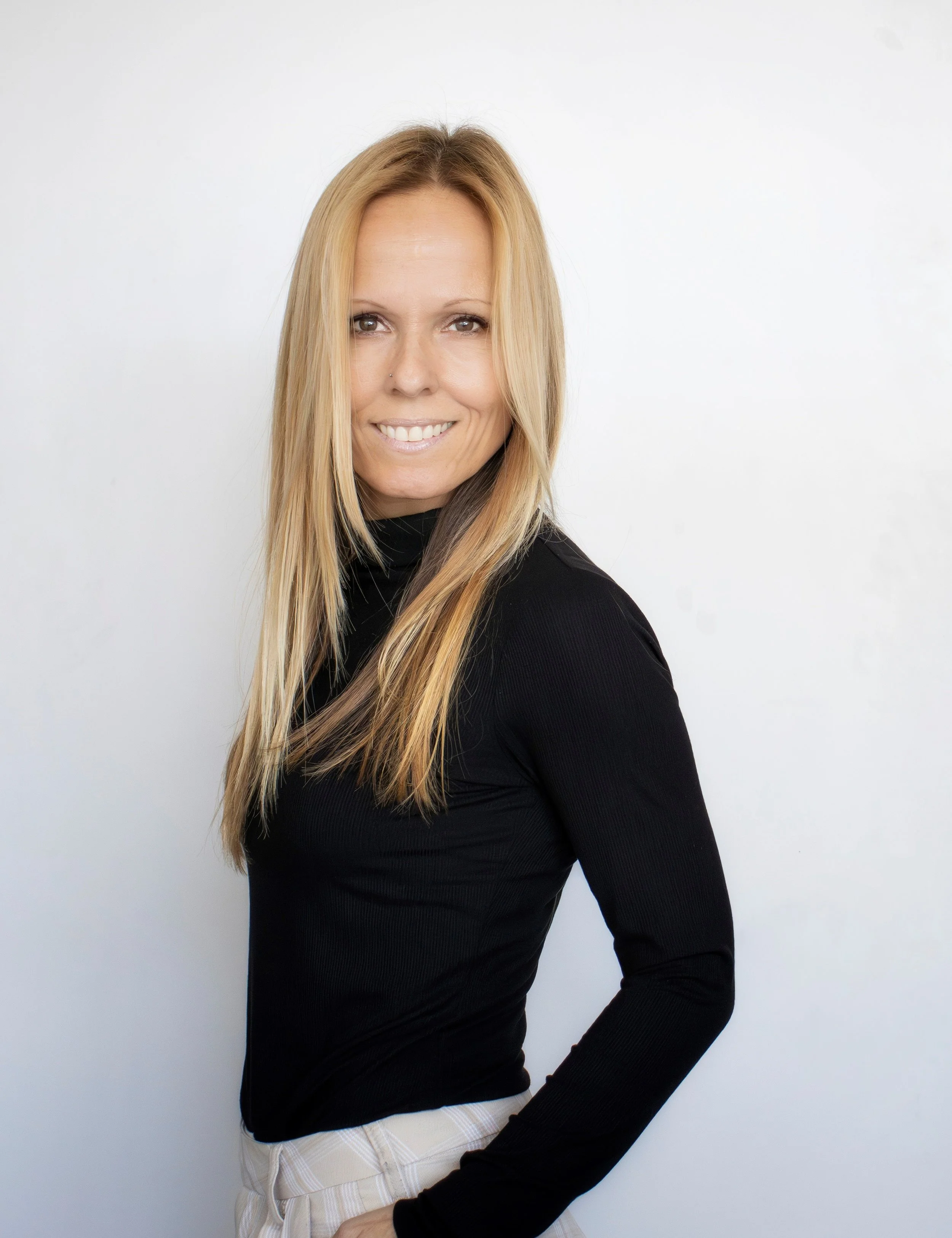 Lächelnde Frau mit langen blonden Haaren, trägt schwarzen Rollkragenpullover und helle Hosen, steht vor weißem Hintergrund.