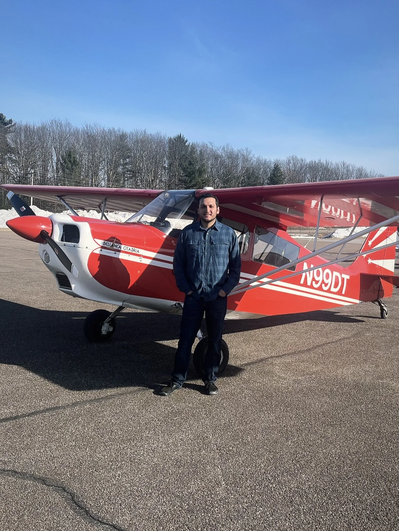 A man standing in front of a red and white small airplane on an airstrip with trees and a clear blue sky in the background.