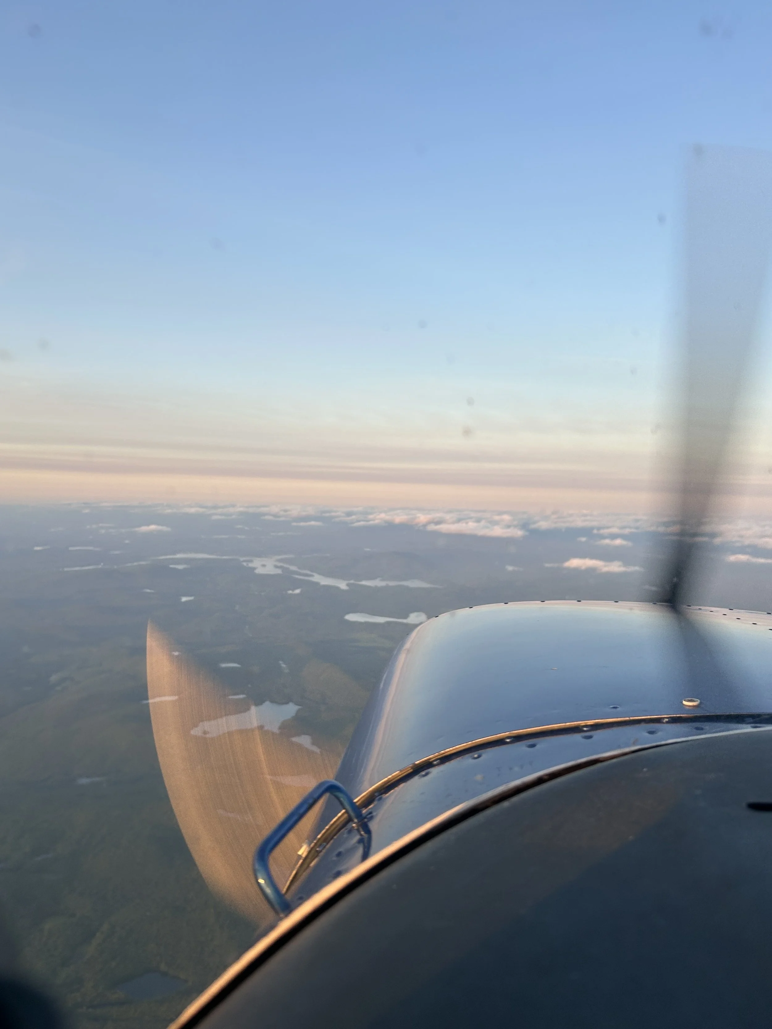 View from inside an airplane cockpit showing the aircraft's wing and a landscape with lakes and green fields below, under a clear blue sky.