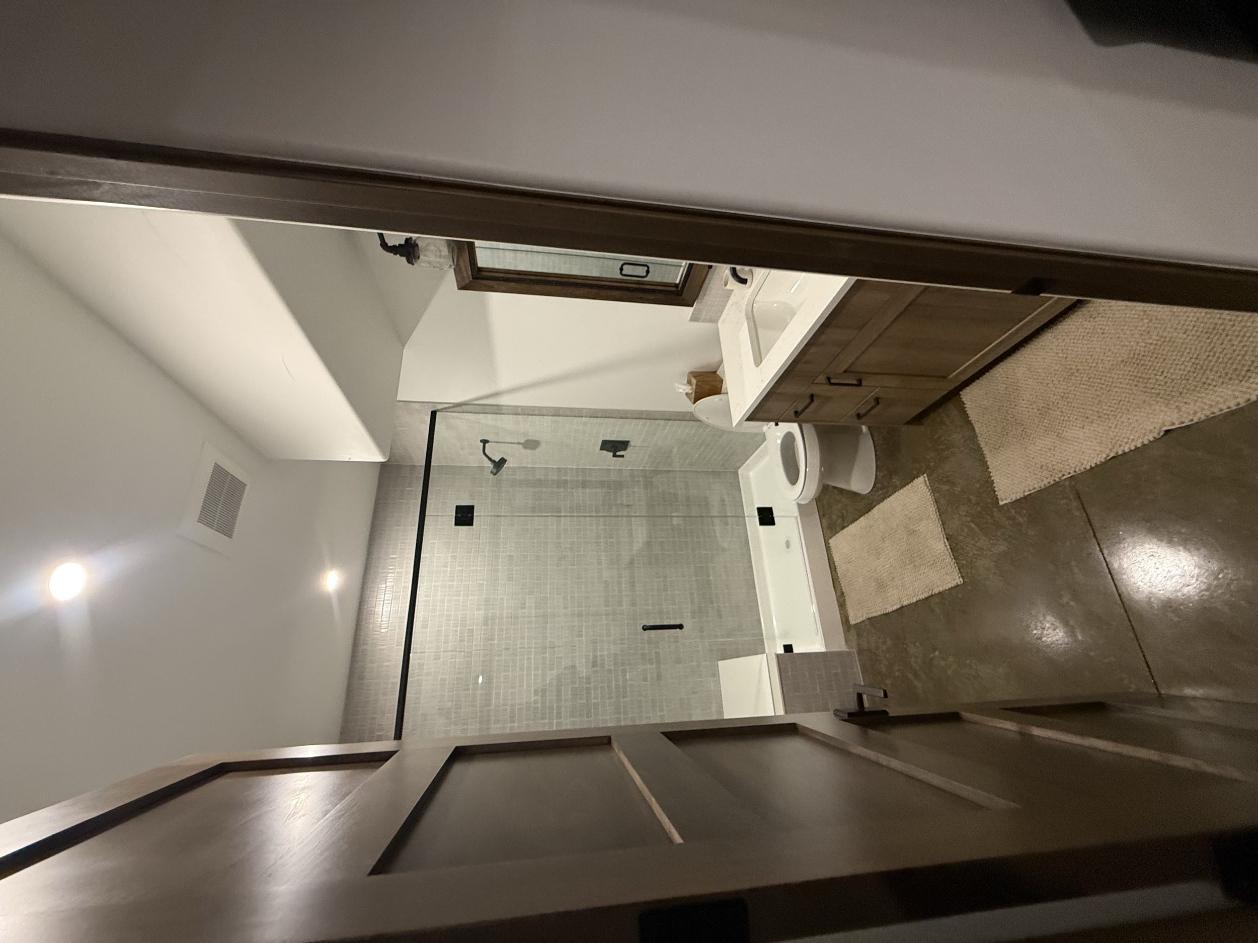 View into a bathroom showing a wooden vanity with a sink, a toilet, a beige rug, and a shower with glass door, beige tiles, and a black showerhead.