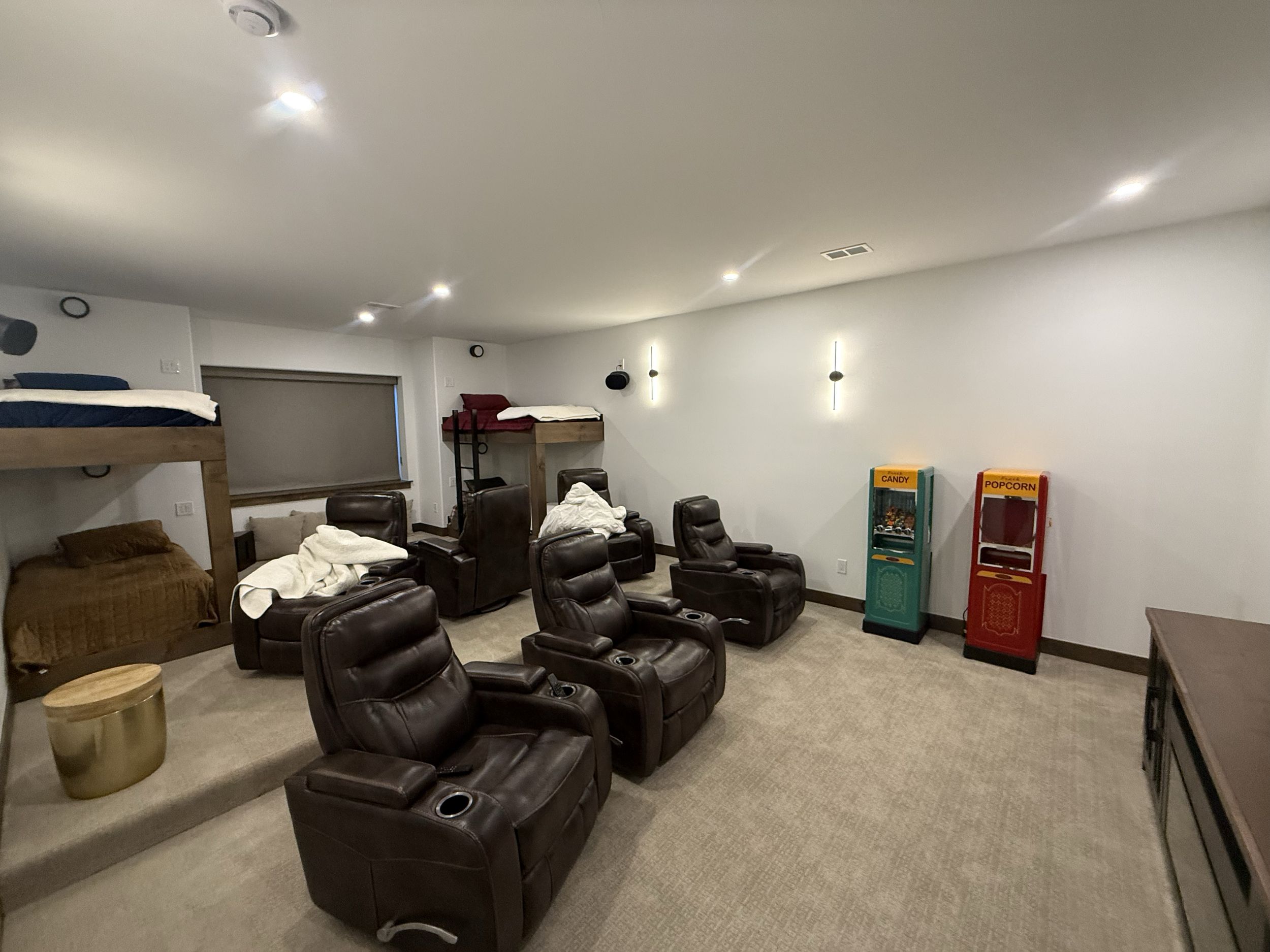A basement room with six brown leather recliners arranged in a circle, some with white blankets. There are two murals, one with two bunk beds on the left and another with a single bunk bed on the right. On the right wall, there are two vintage-style 