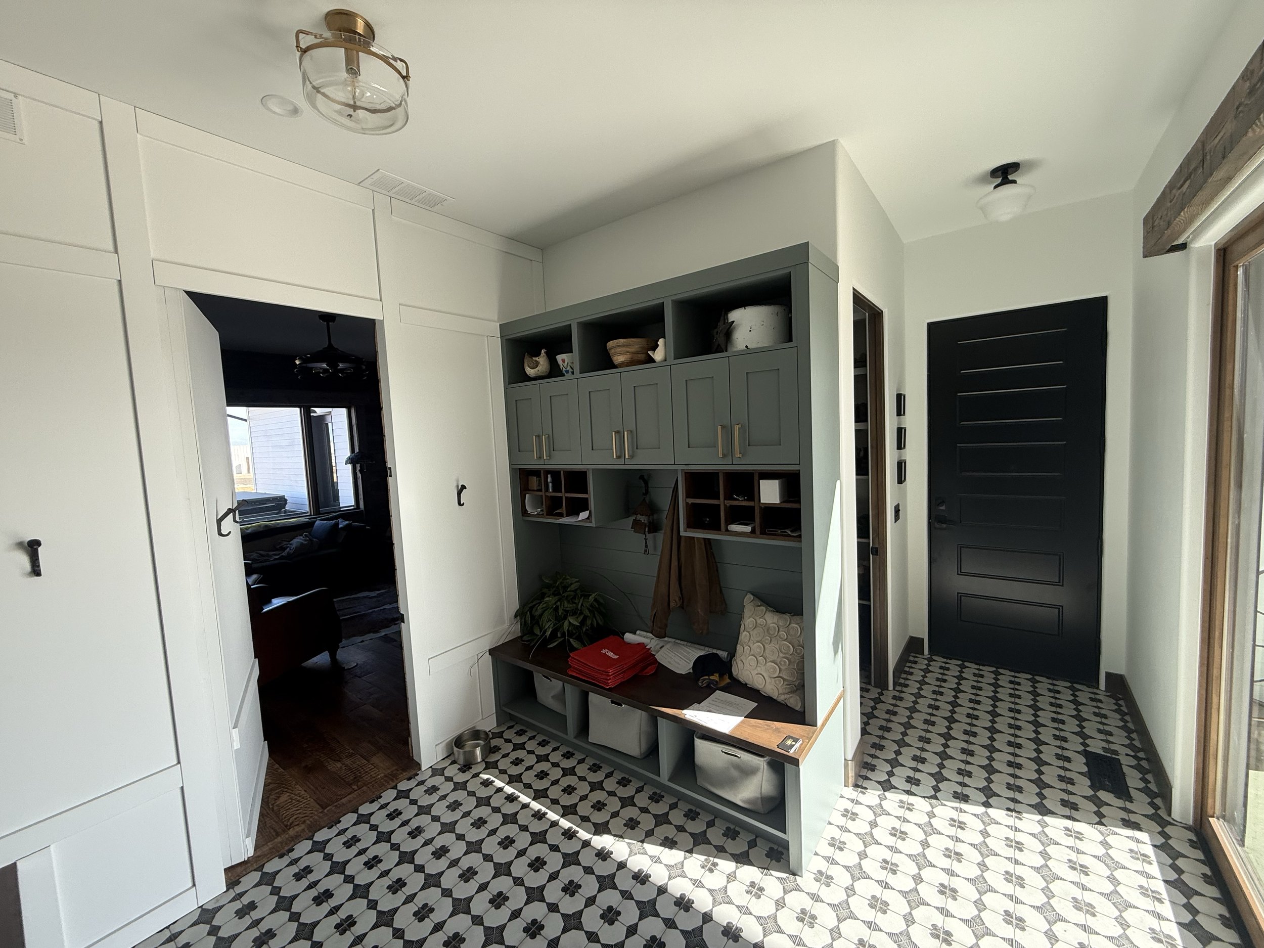 Entryway with black front door, built-in blue cabinet with cubbies, black and white patterned tile floor, large window, white walls, and visible sunlight.
