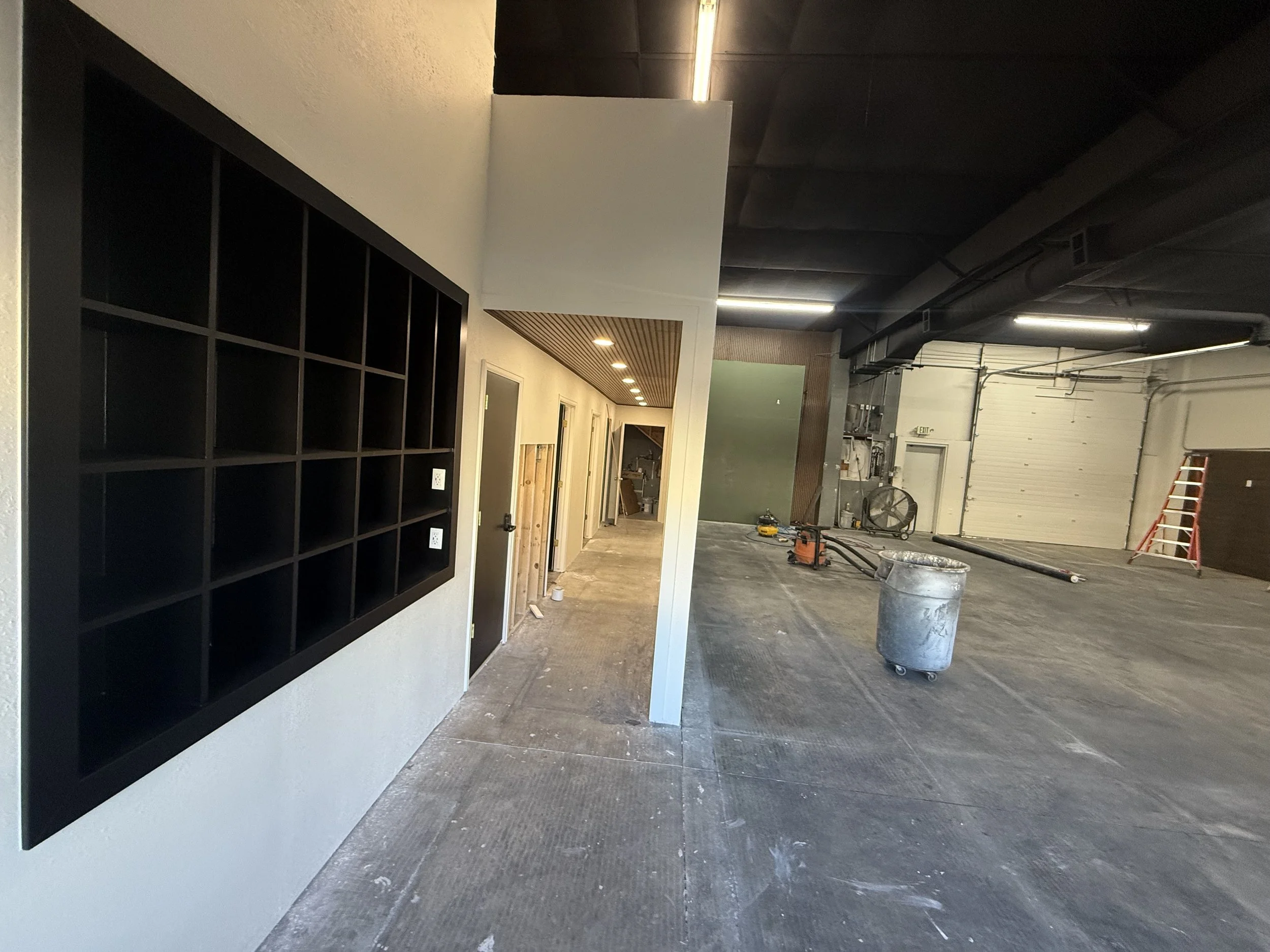 Indoor construction site with a black grid shelving unit on the left wall and unfinished flooring; construction tools and equipment visible in the background.