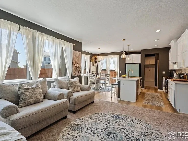 A spacious, open-concept living room and kitchen with large windows, white curtains, beige sofas, a carpet, and a dining area with a table and chairs. The kitchen features white cabinets, a kitchen island with a yellow faucet, and modern appliances.