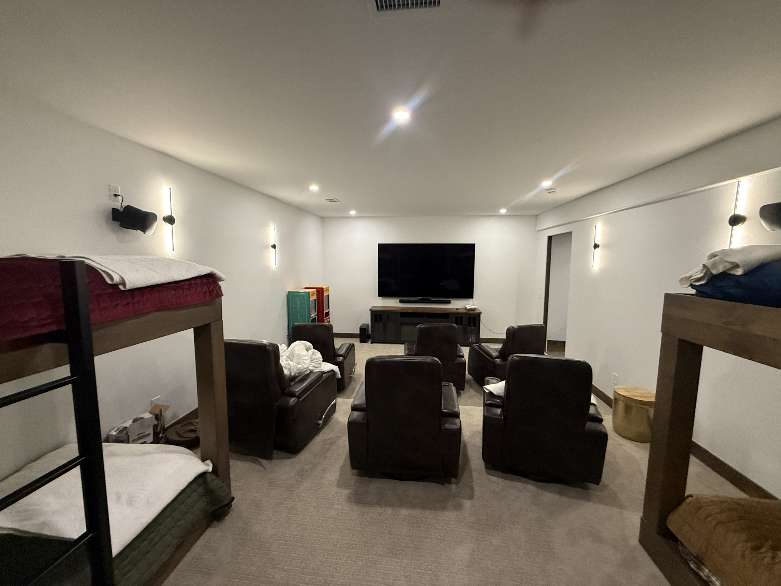 A spacious living room with a large flat-screen TV mounted on the wall, surrounded by several brown armchairs arranged for viewing. There are bunk beds on each side of the room, one with a red blanket and the other with a blue blanket. The walls have