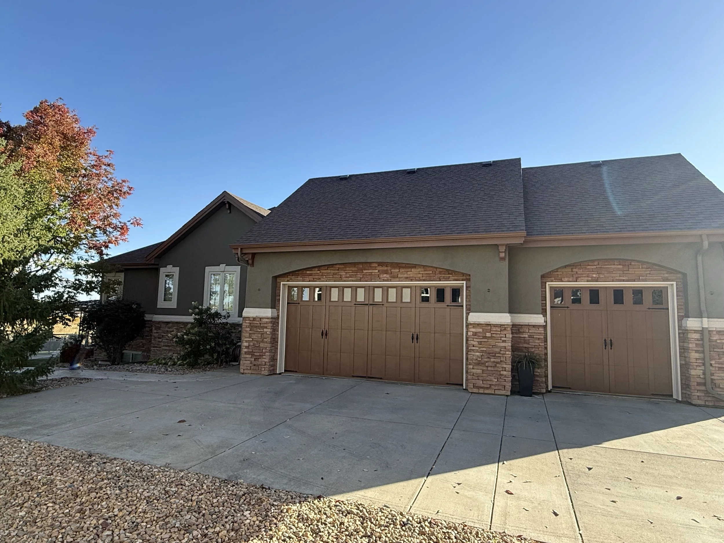 A modern house with double garage doors, brick and stucco exterior, a front driveway, and landscaped side yard with small trees and shrubs. Clear blue sky in the background.