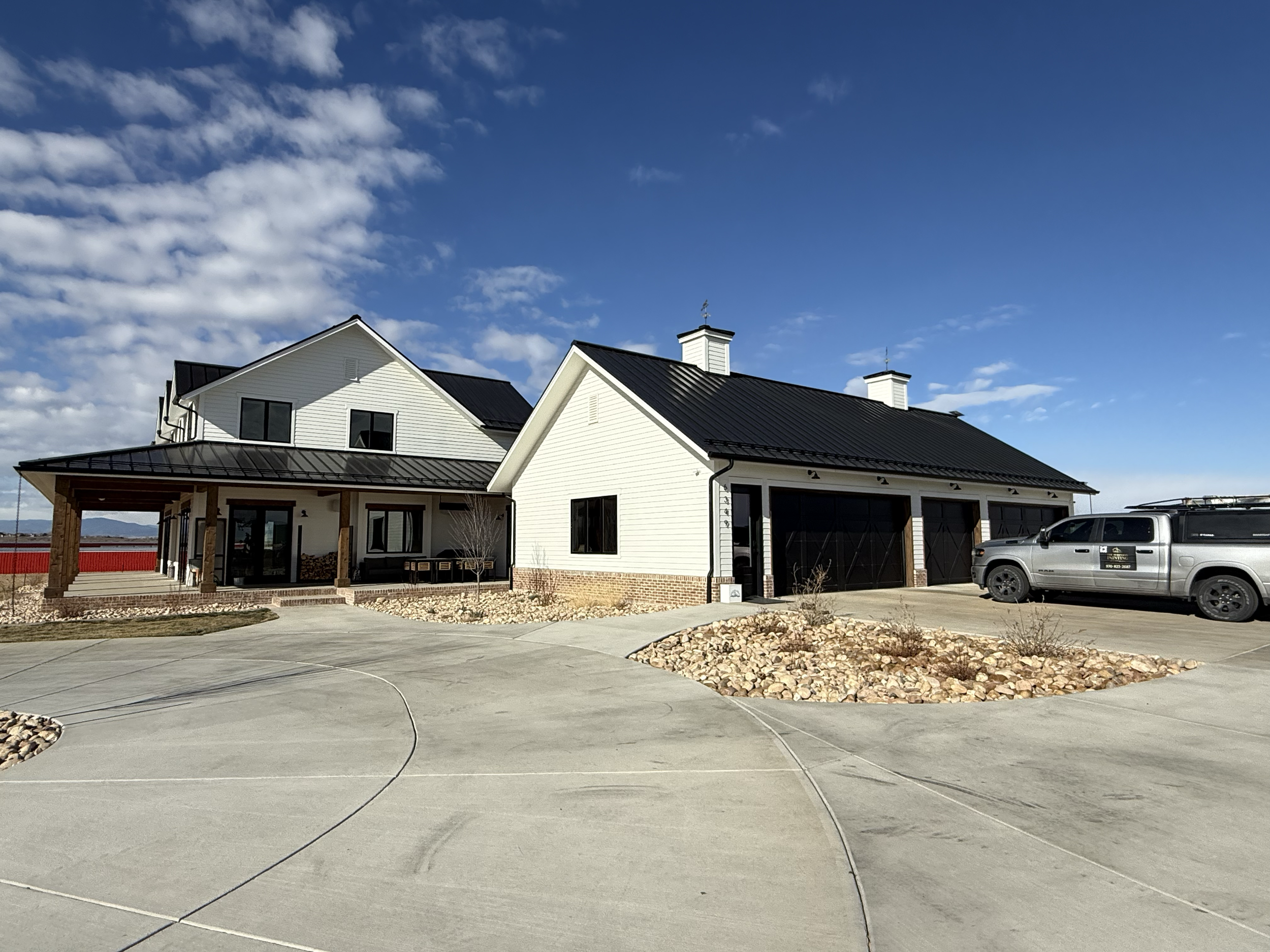Modern white house with black metal roof, two stories, and attached garage, parked black truck, concrete driveway, landscaped with rocks and small plants, under blue sky with clouds.