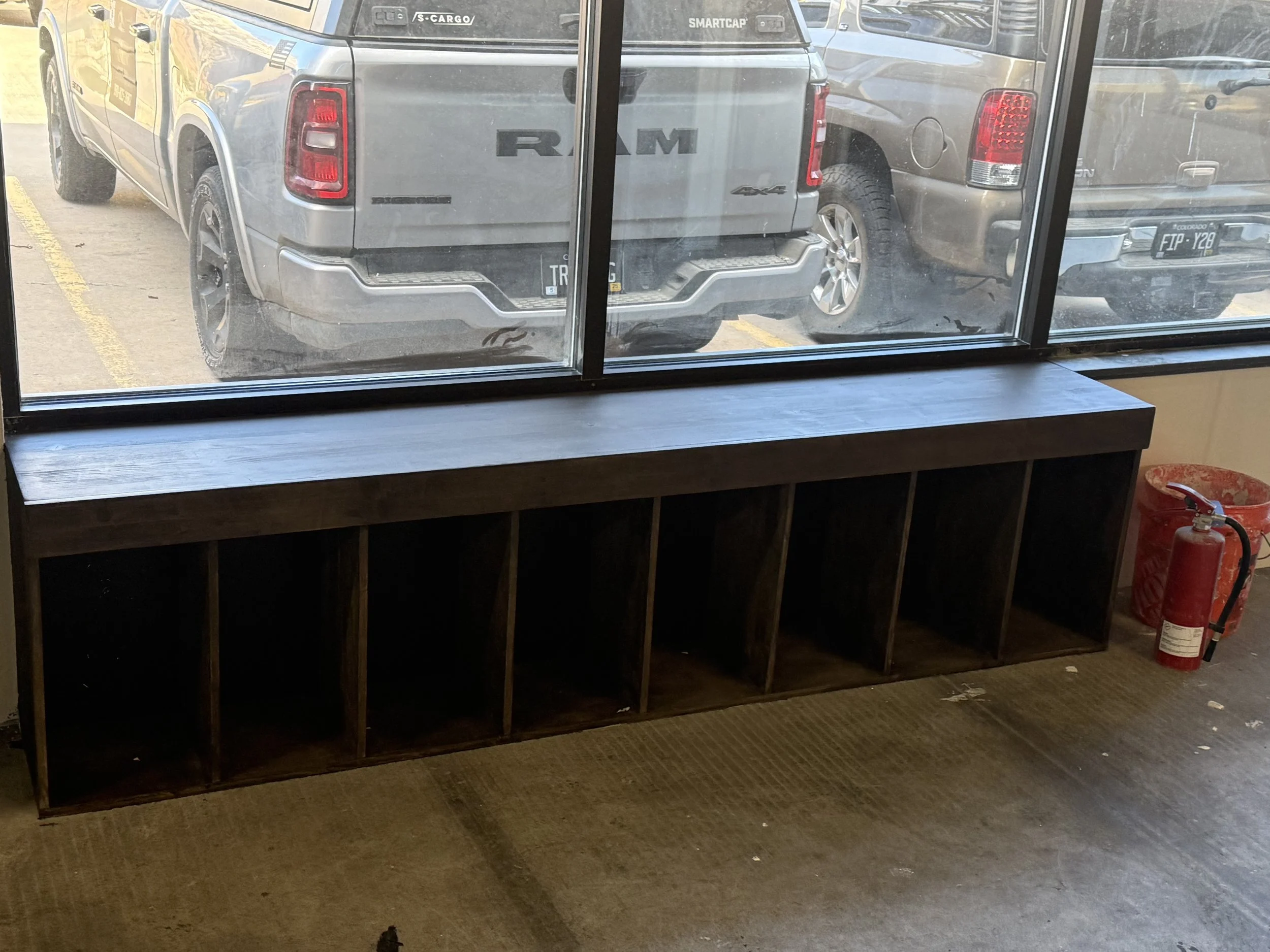 Empty wooden cubby shelf near a large window with parked trucks outside, a fire extinguisher and trash can to the side.