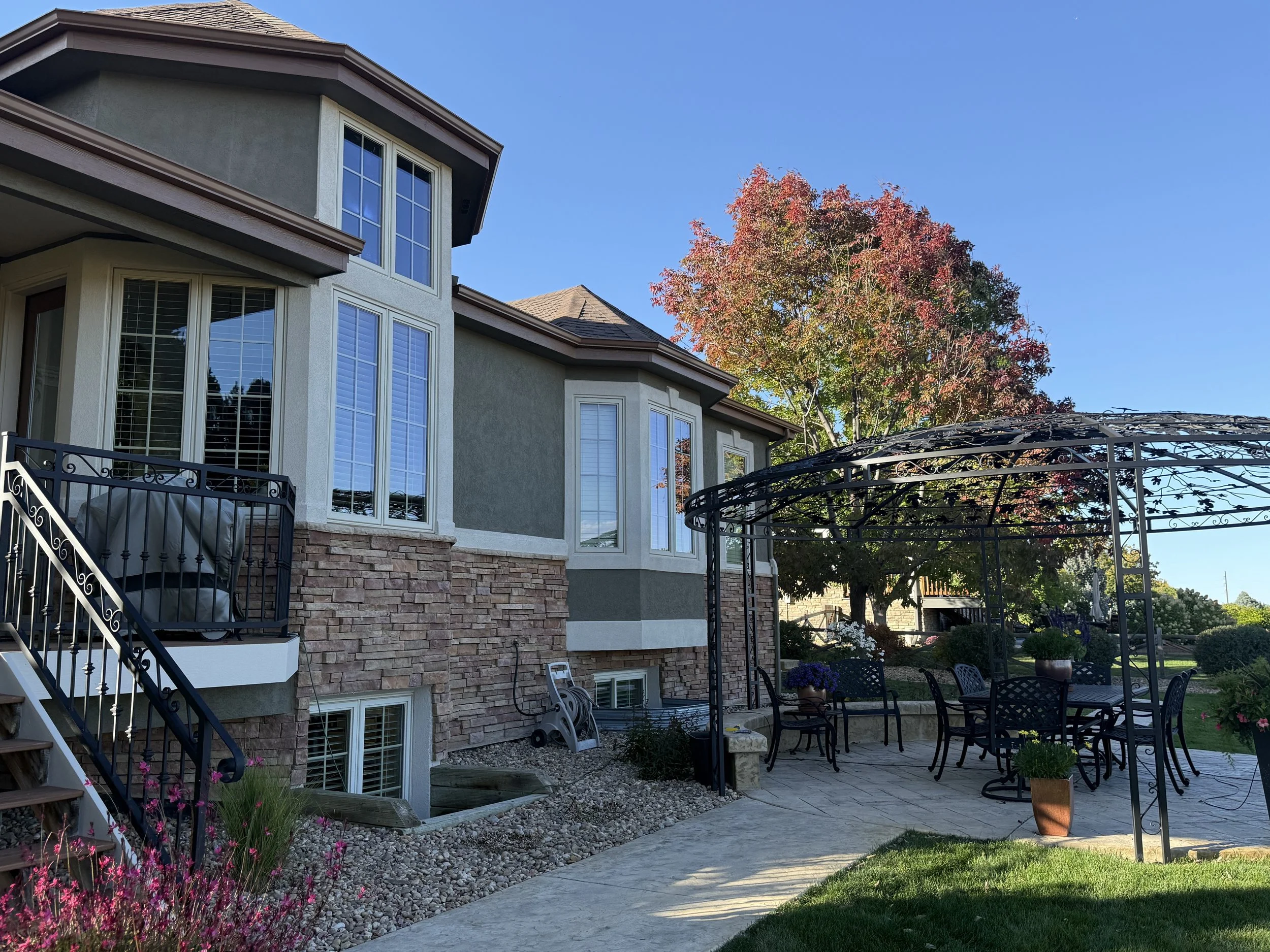 Exterior view of a two-story house with multiple white windows, a small black dining table and chairs under a metal gazebo, a large tree with red and green leaves, and a clear blue sky.