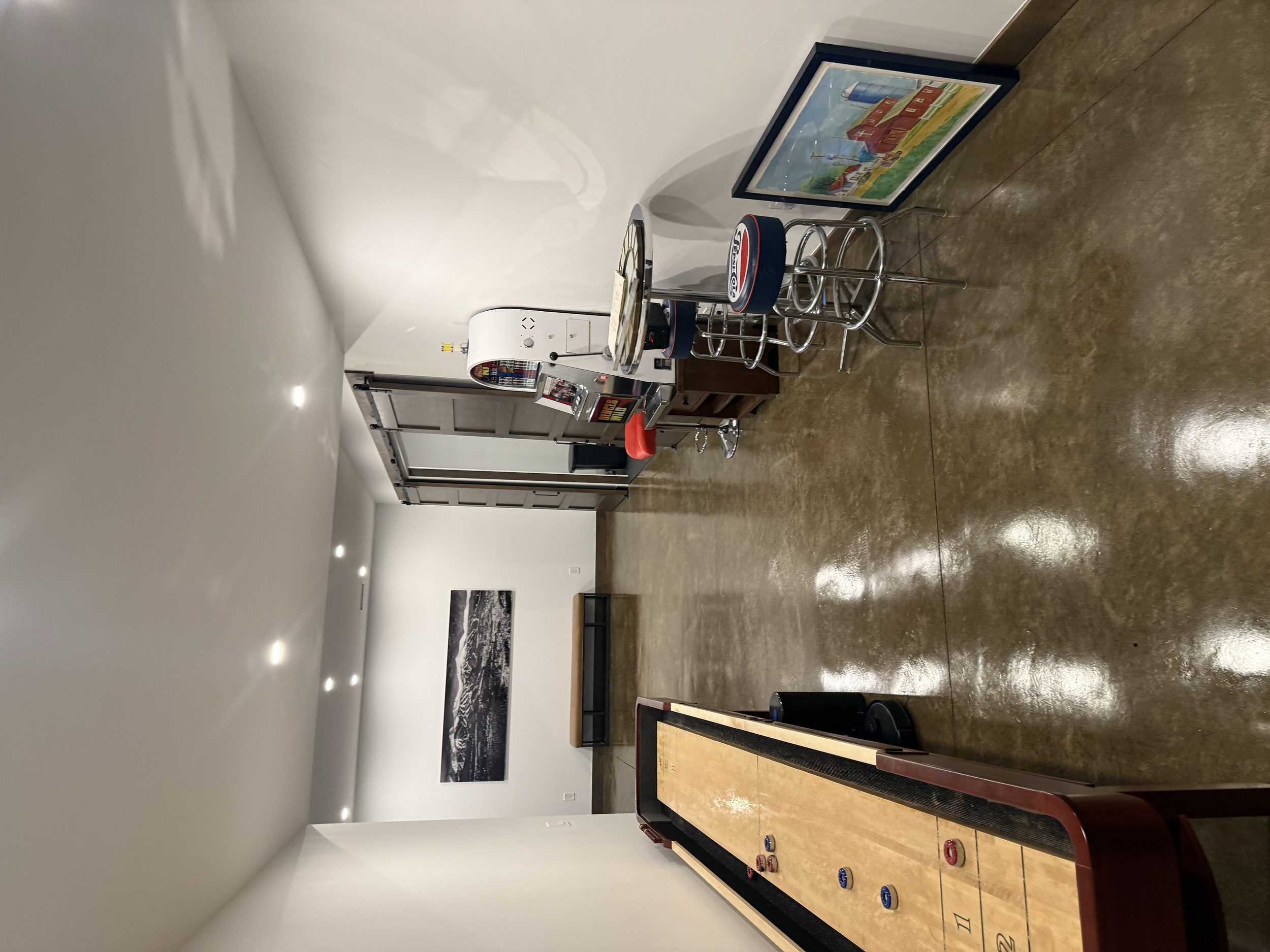 Indoor space with a shuffleboard table, framed artwork on the wall, and a seating area with stools and a countertop in the background.