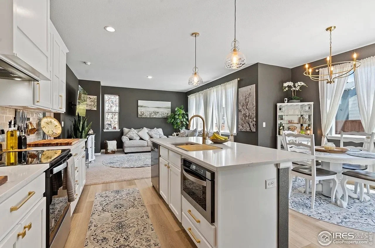 Open-concept living room and kitchen with white cabinets, a kitchen island, and dining area, featuring dark gray walls, white furniture, and large windows with white curtains.