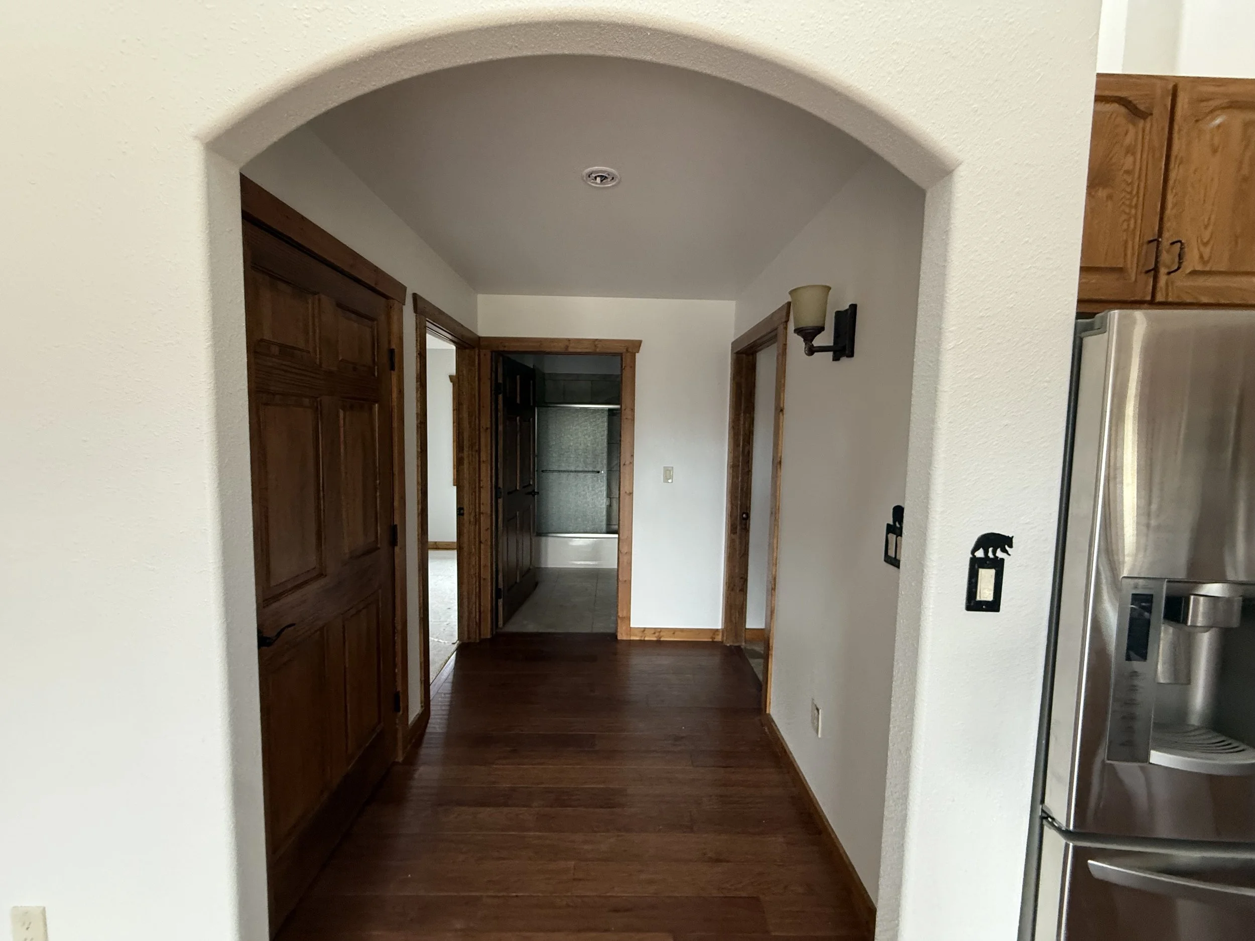View of a hallway with wooden floors and doors, leading to a bathroom with a bathtub, inside a house with cream-colored walls and wood trim. Part of a stainless steel refrigerator is visible on the right.