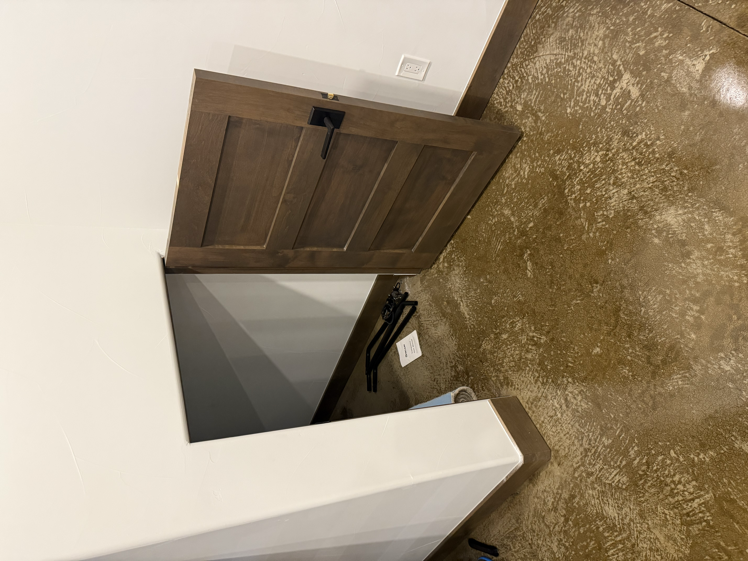 An interior room with a brown wooden door, brown trim, a white wall, a wall outlet, and a brown textured floor. There are some black metal objects and a small piece of paper on the floor.