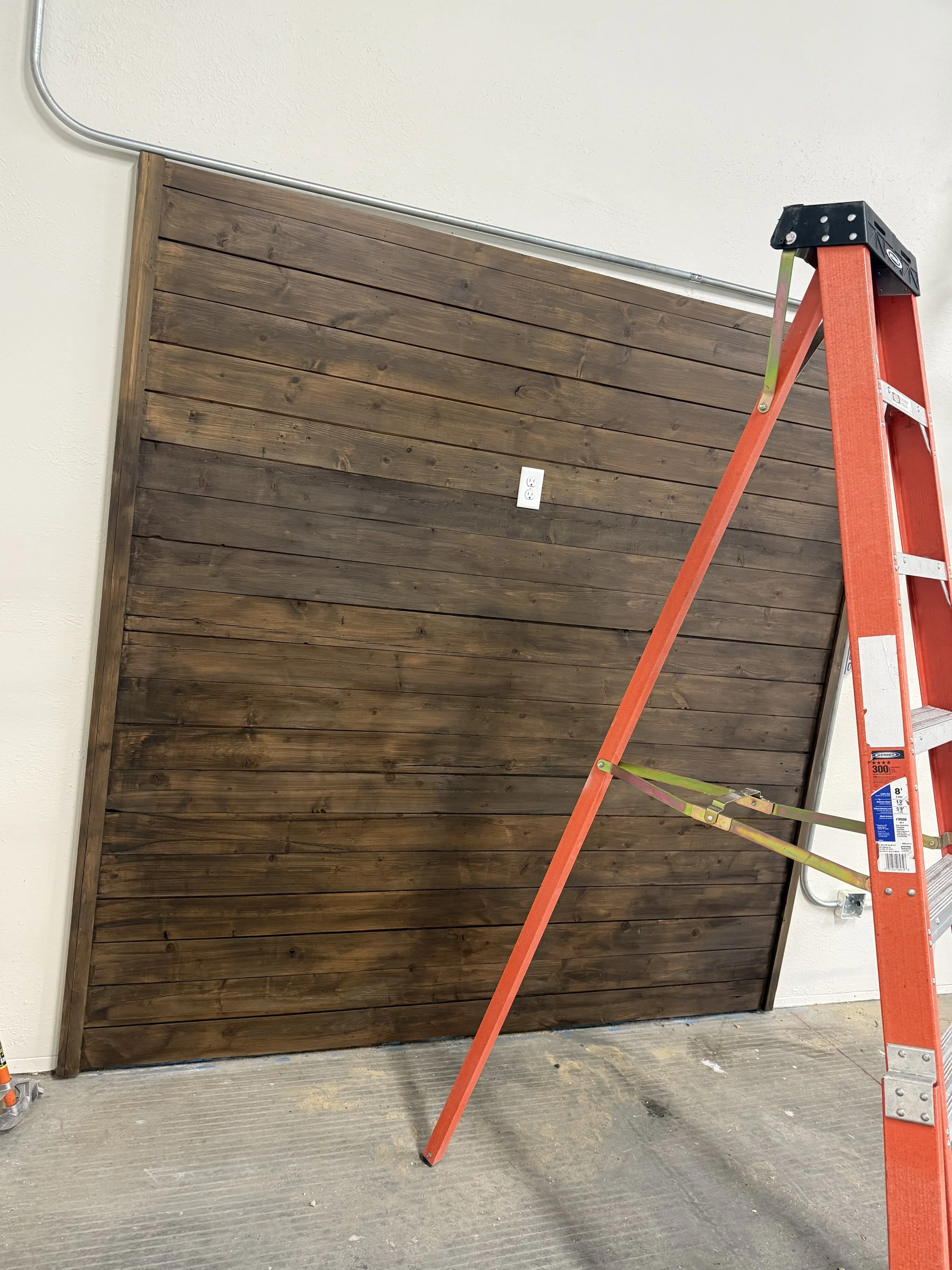 Interior wall with wooden paneling, a ladder leaning against it, with a single electrical outlet in the center of the wall.