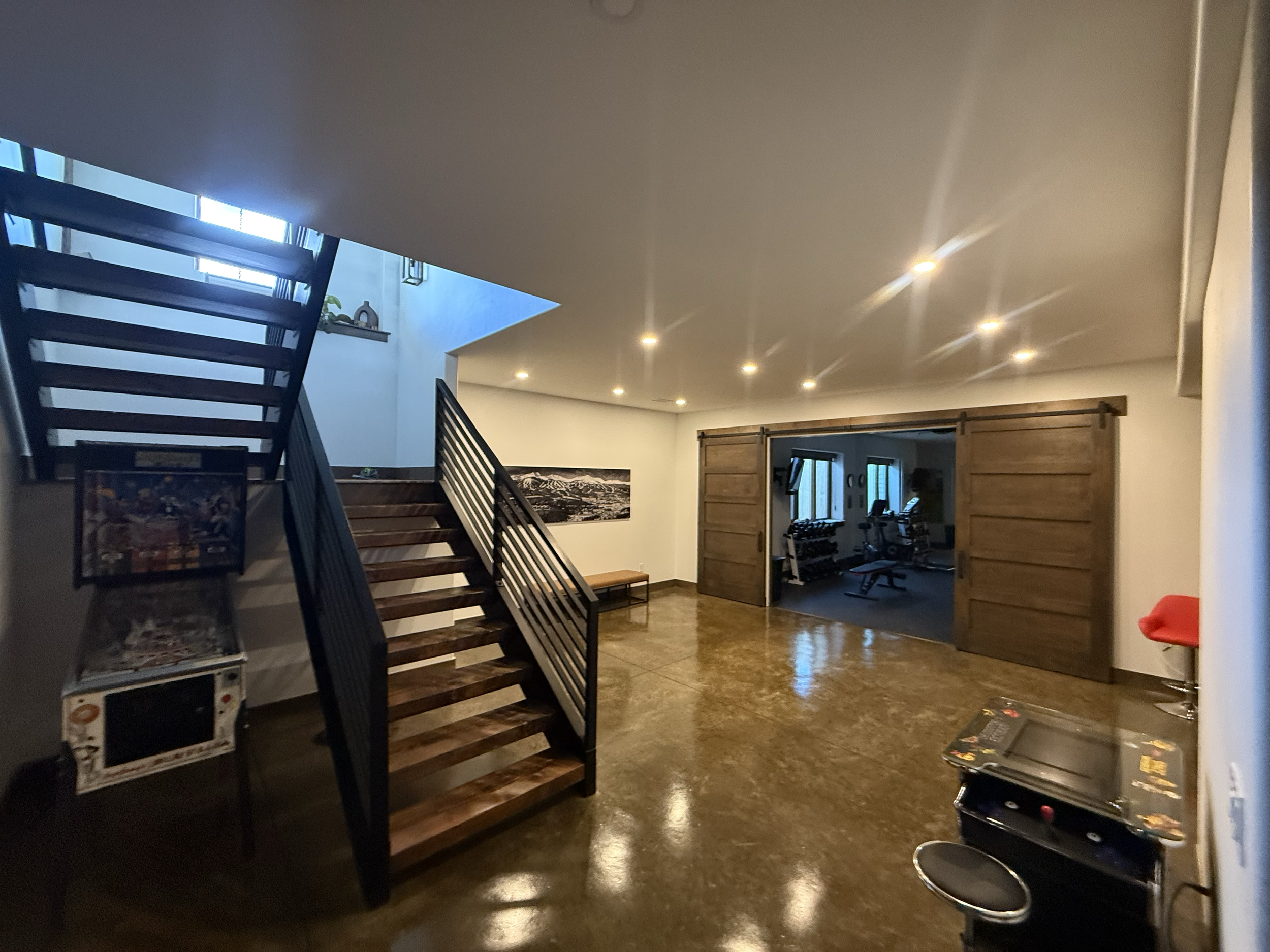 Interior view of a house with a staircase, a wooden door, artwork on the wall, and a room with exercise equipment, with shiny polished floors and overhead lighting.