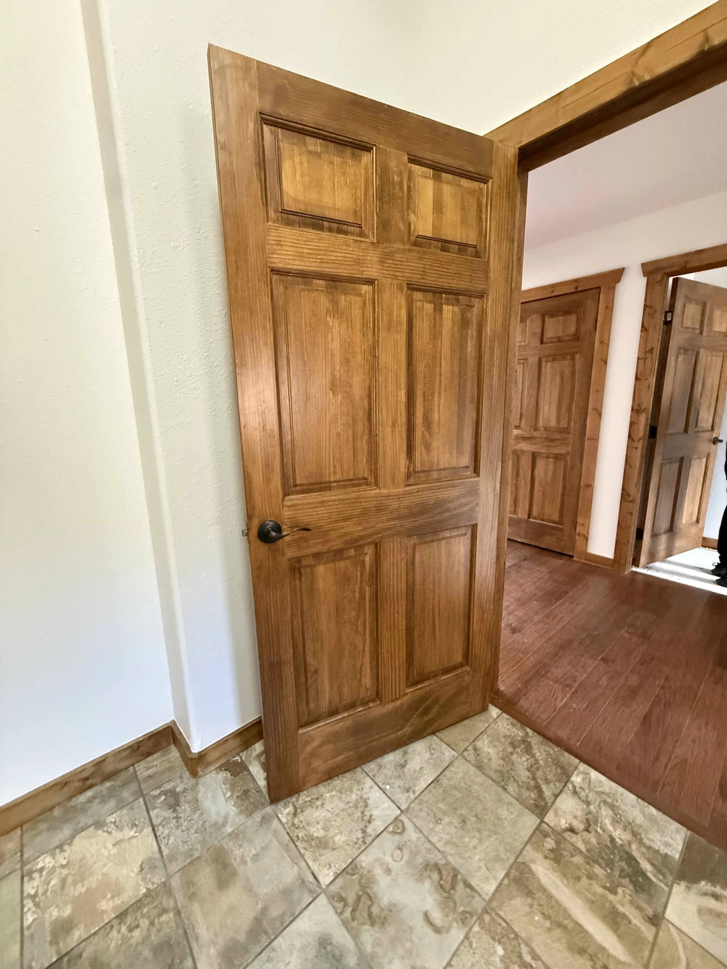 View of a wooden door ajar, showing multiple interior doors in a home with wood trim, tiled and hardwood floors.
