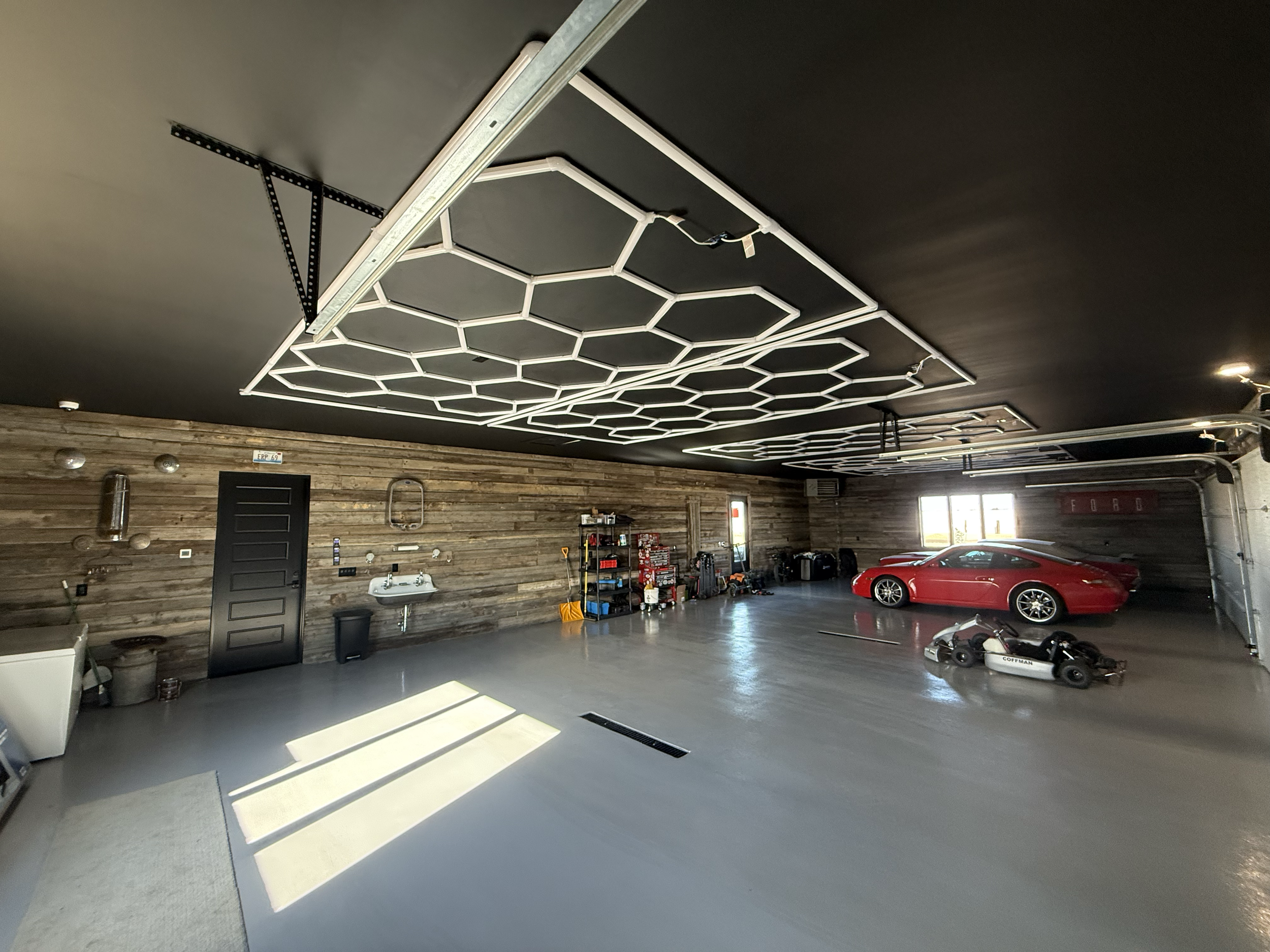 A spacious garage with a black ceiling featuring white geometric LED light fixtures, wooden paneled walls, and a smooth concrete floor. The garage contains a red sports car, a small go-kart, and various tools and storage shelves along the walls.
