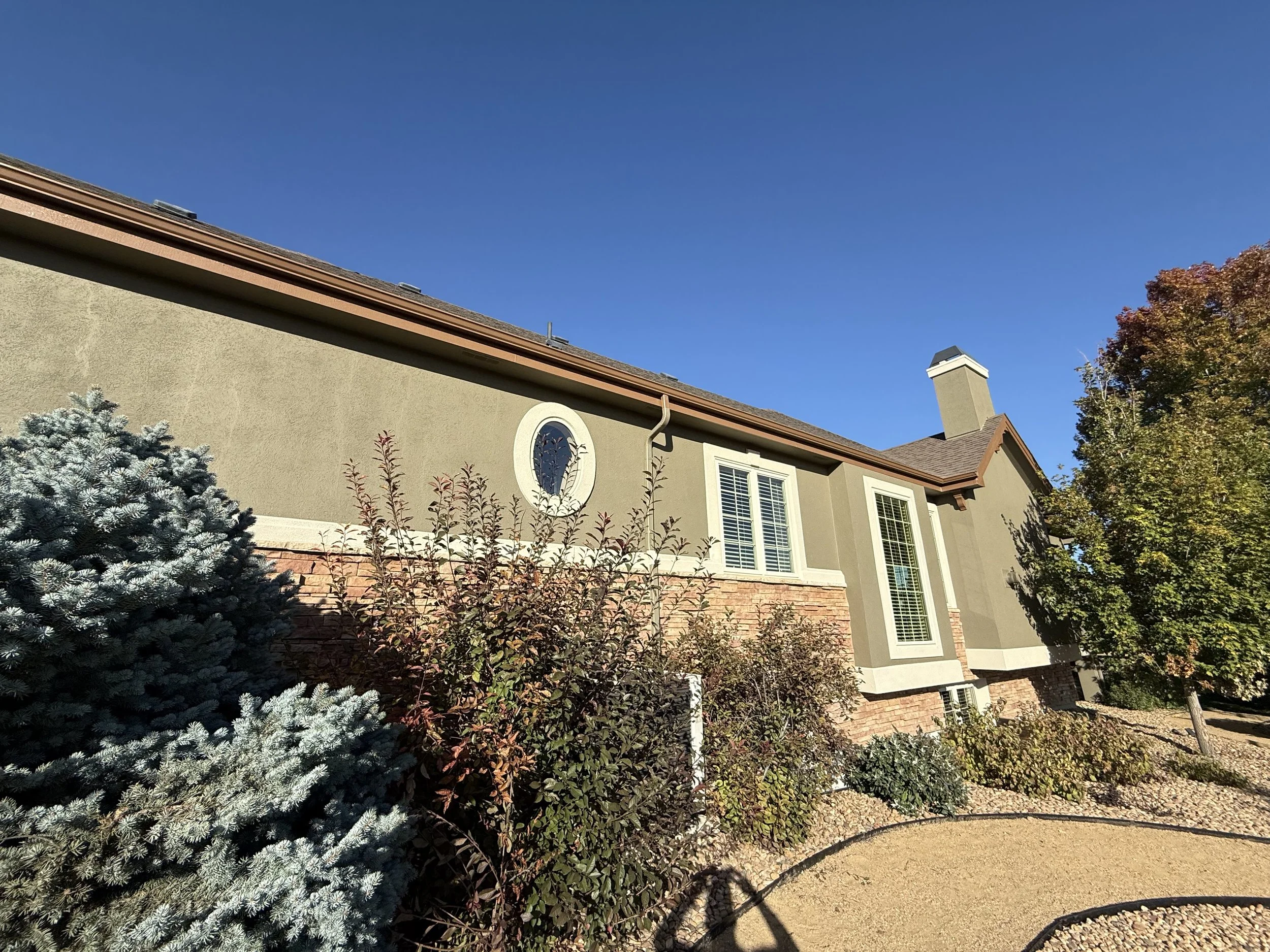 Exterior of a house with beige walls, brick accents, multiple windows with white shutters, and a tall chimney, surrounded by trees and bushes under a clear blue sky.