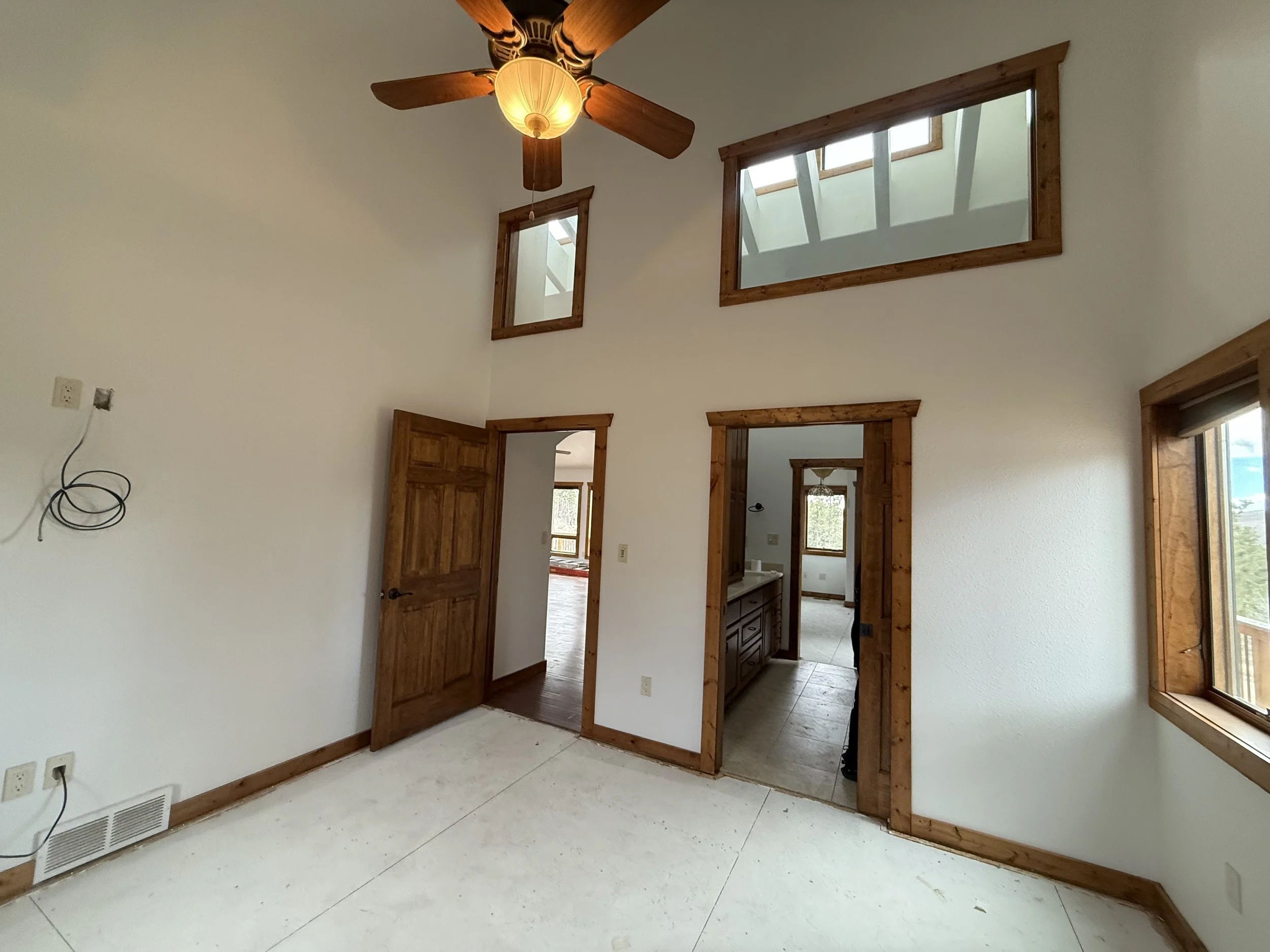 Empty room with white walls, wooden trim, two windows on a high sloped wall, one window on the right, and a ceiling fan with a light. The room has an open doorway leading to a bathroom and another opening leading to an adjacent room.